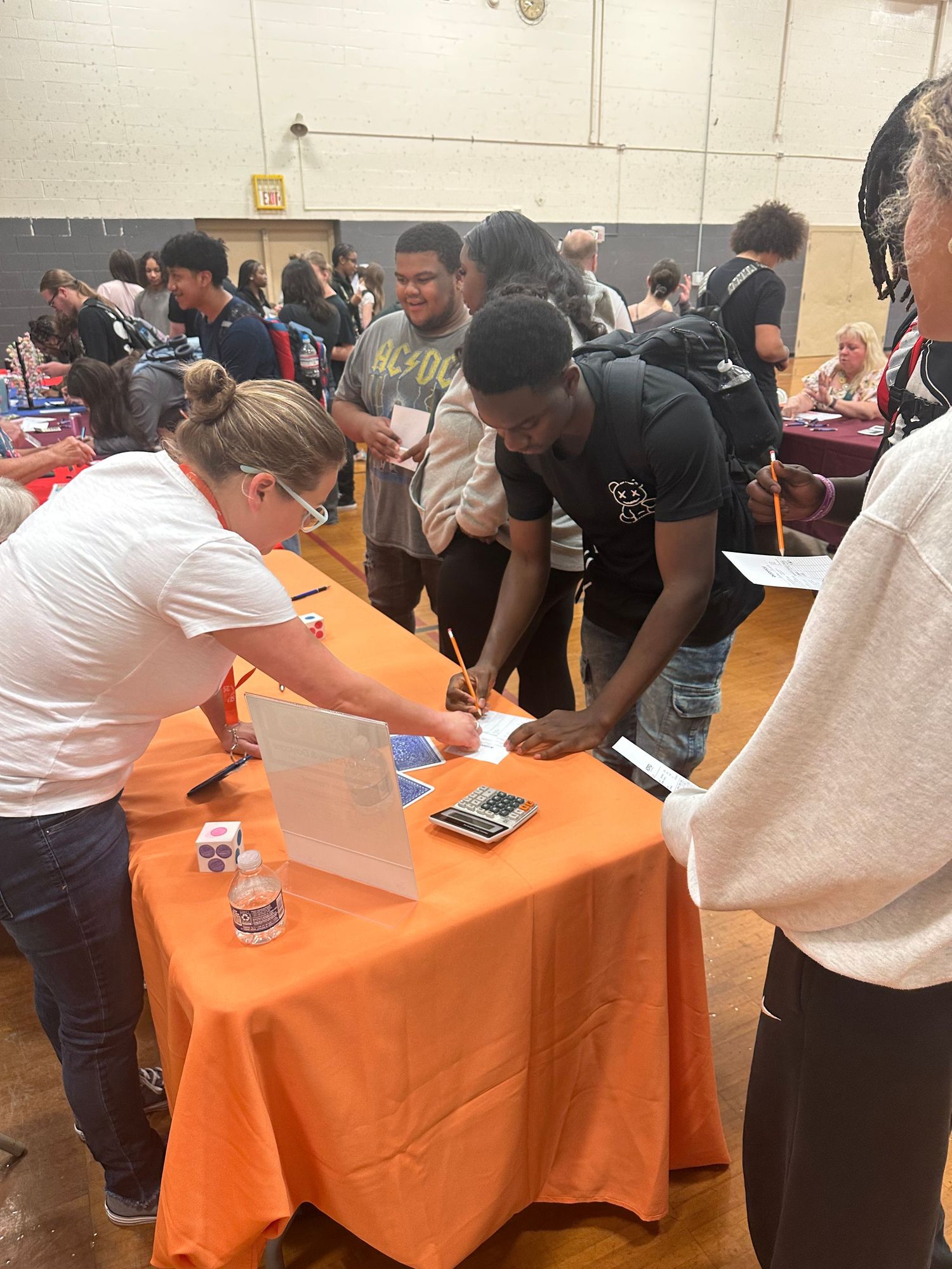 Students signing up at an orange-draped table during a school career fair in a gymnasium.