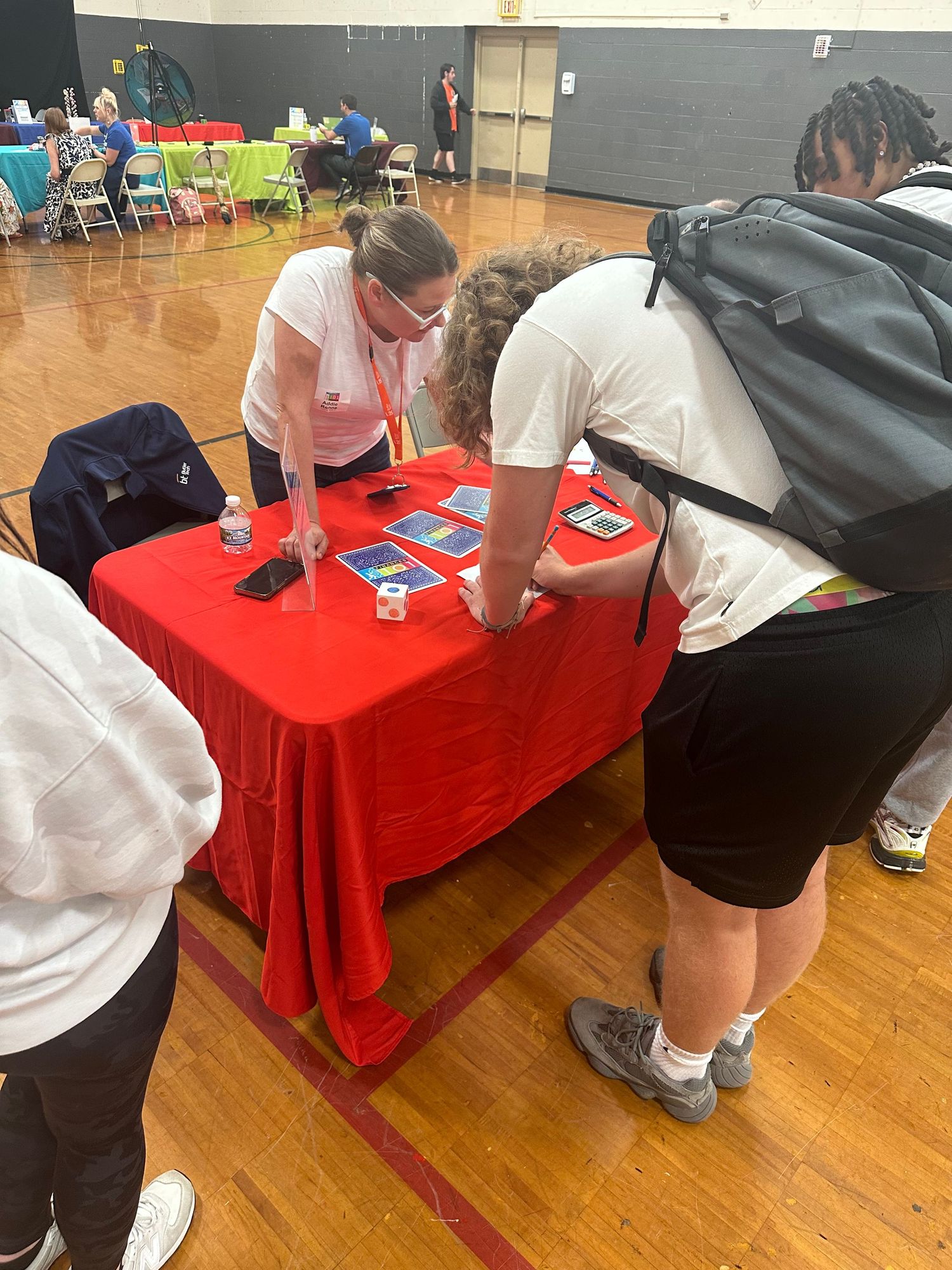 Students gather around a red table with tablets during an indoor educational fair in a gymnasium.