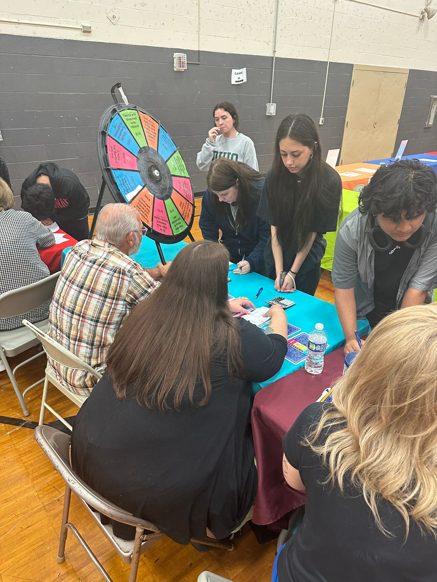 Students and adults gather around a table with a colorful prize wheel at an indoor school fair event.