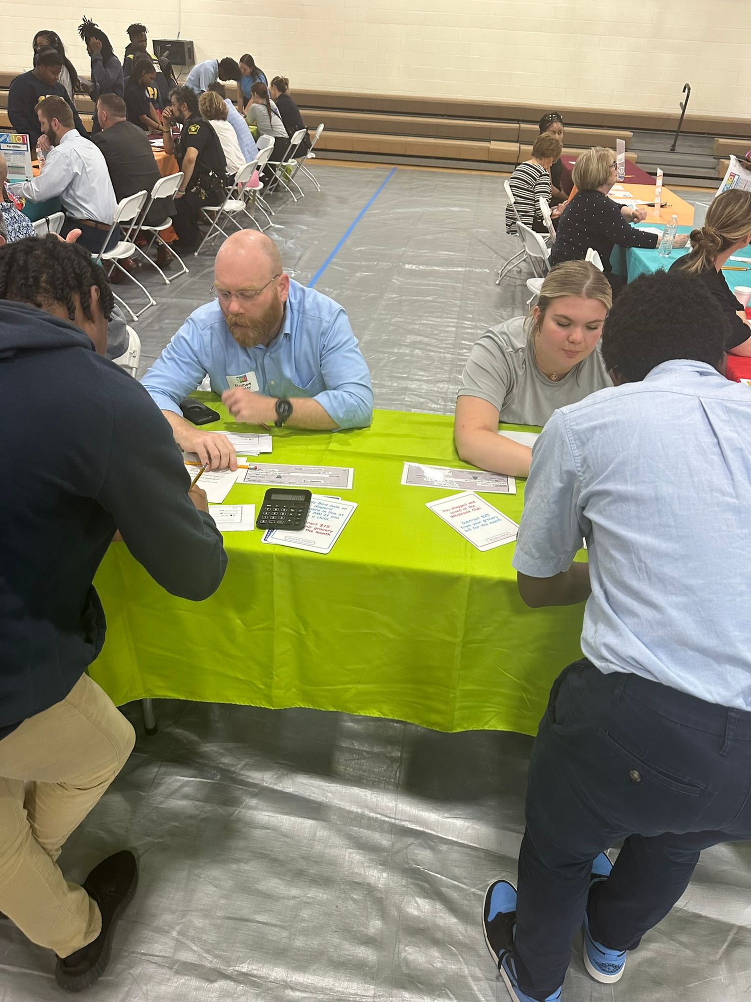 Adults and students collaborate around a green table during an educational event in a school gymnasium.