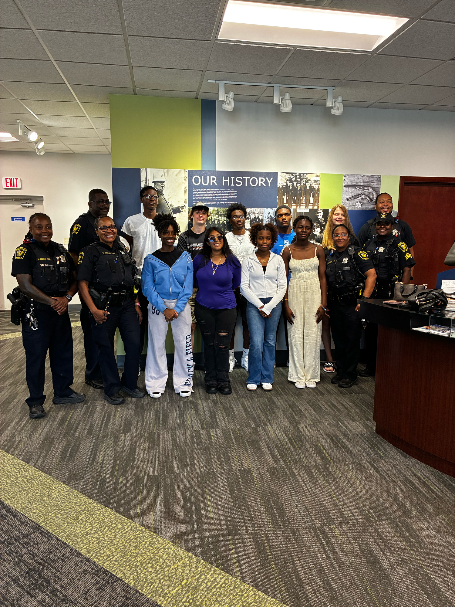 Group of students and police officers posing together indoors in front of an Our History wall display.