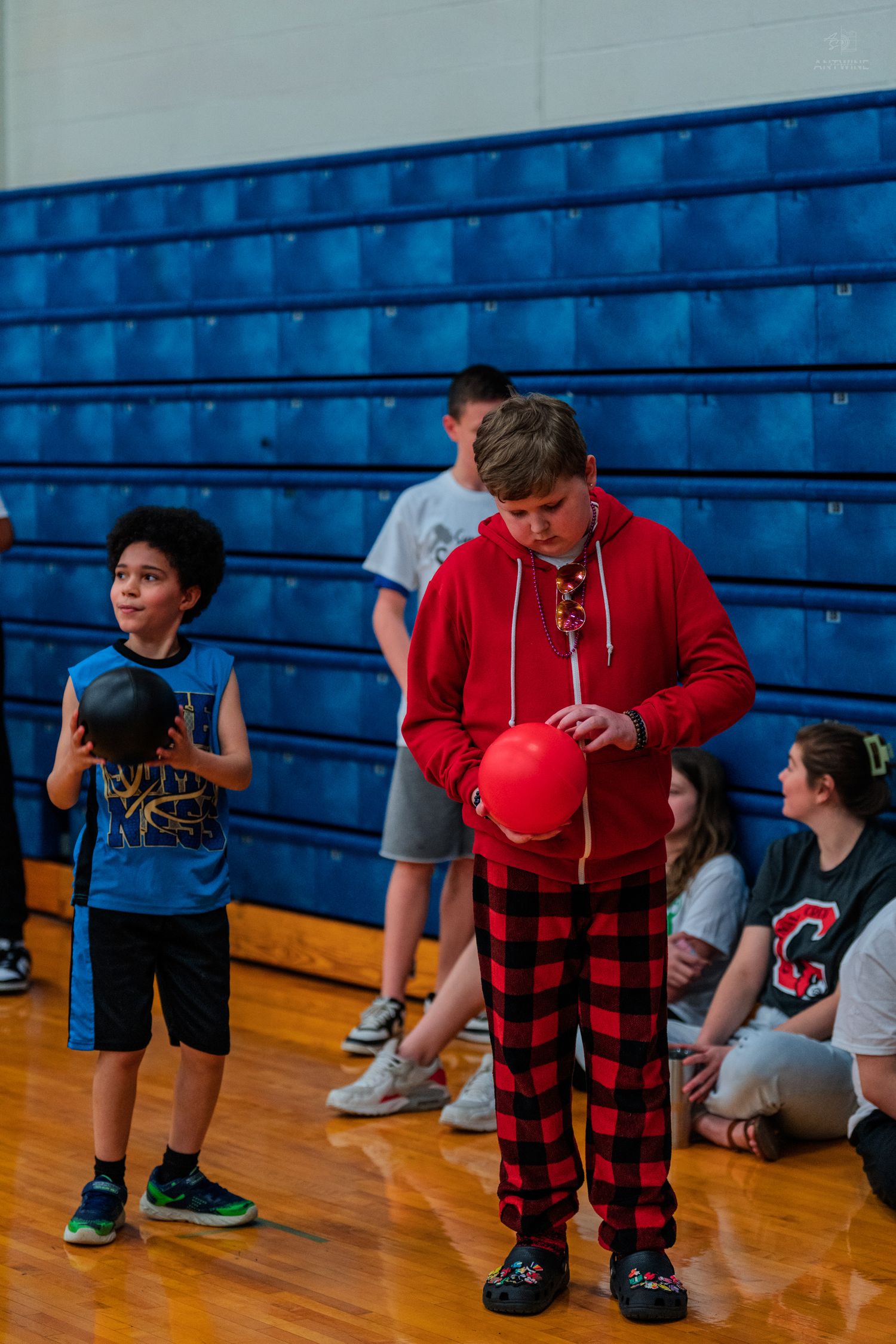 Children playing dodgeball in a school gymnasium, boy in red hoodie and plaid pants holds red ball.