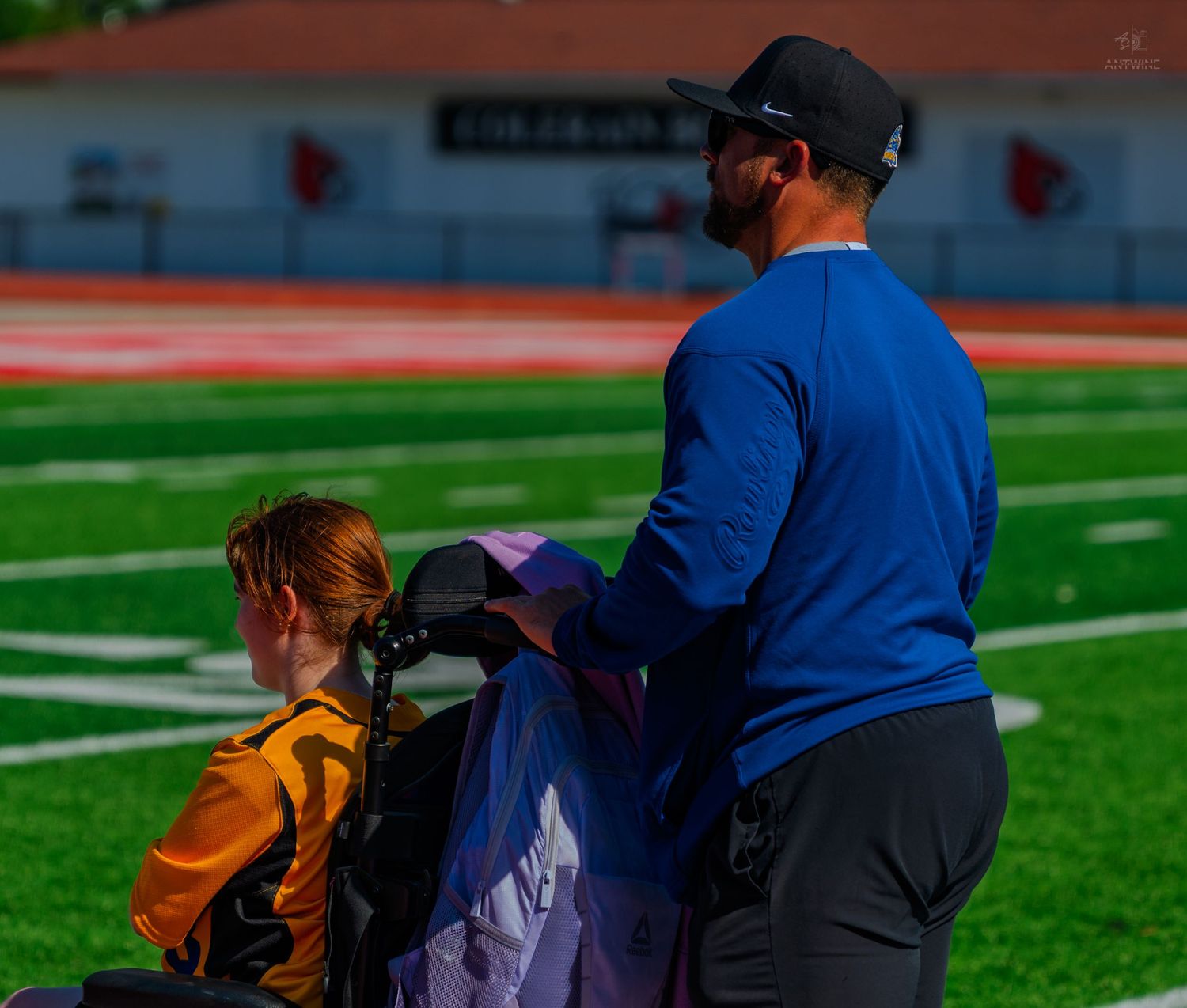 Man in blue shirt assists a young athlete in a wheelchair on a football field with red track.