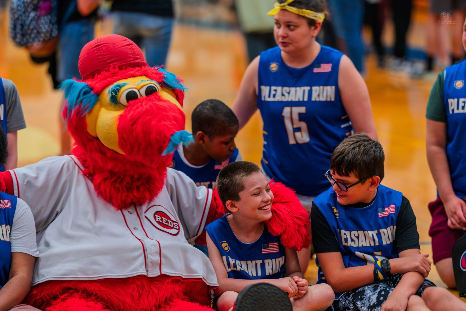 Cincinnati Reds mascot interacts with smiling children in Pleasant Run basketball uniforms at a special needs sports event.