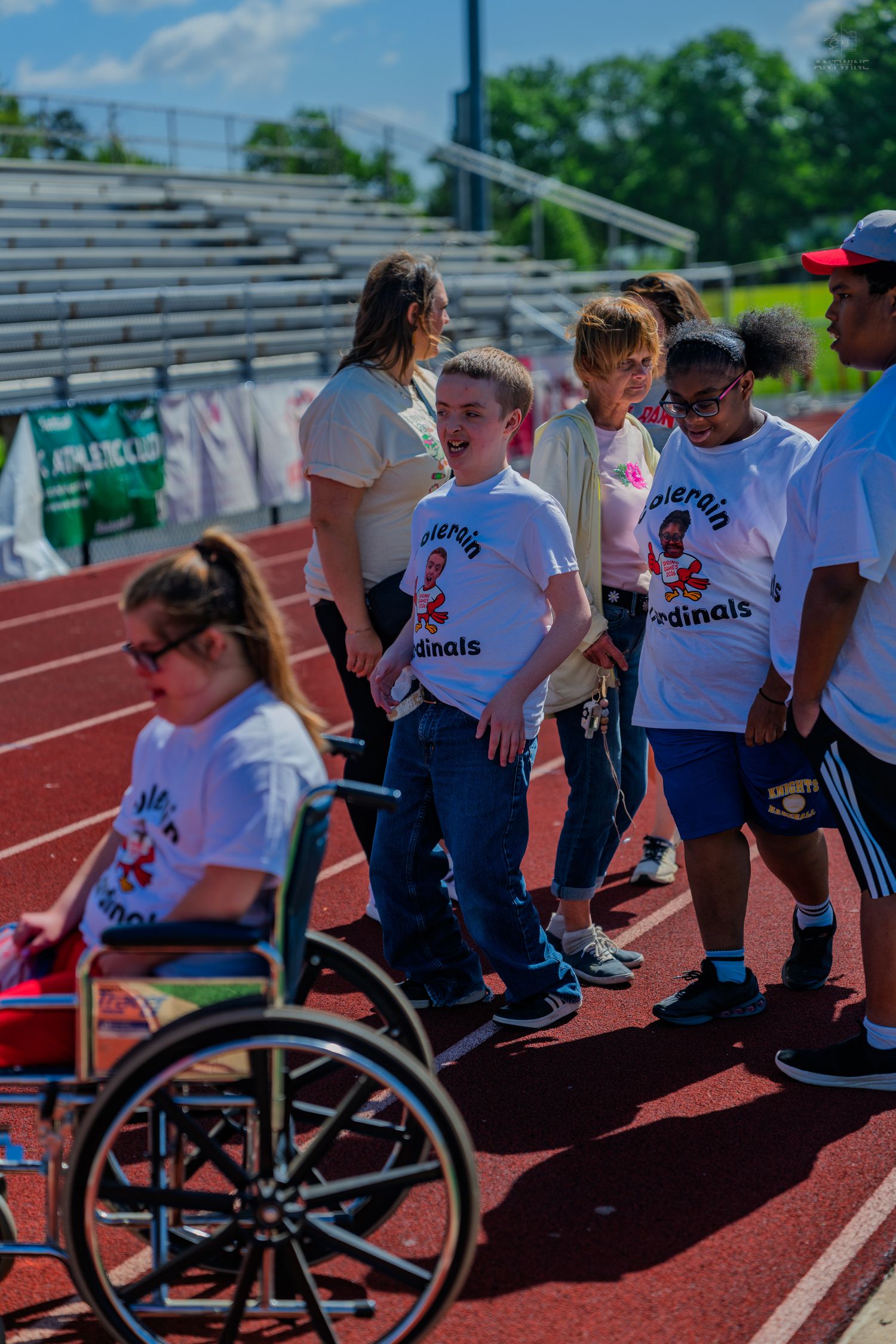 Children in Cardinals shirts stand on a track at a school athletic event, with a wheelchair visible in the foreground.