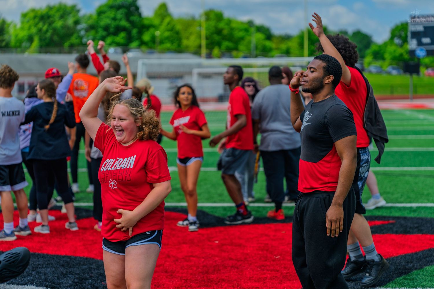 Students in red shirts celebrate on a football field during an outdoor school sports event on a sunny day.