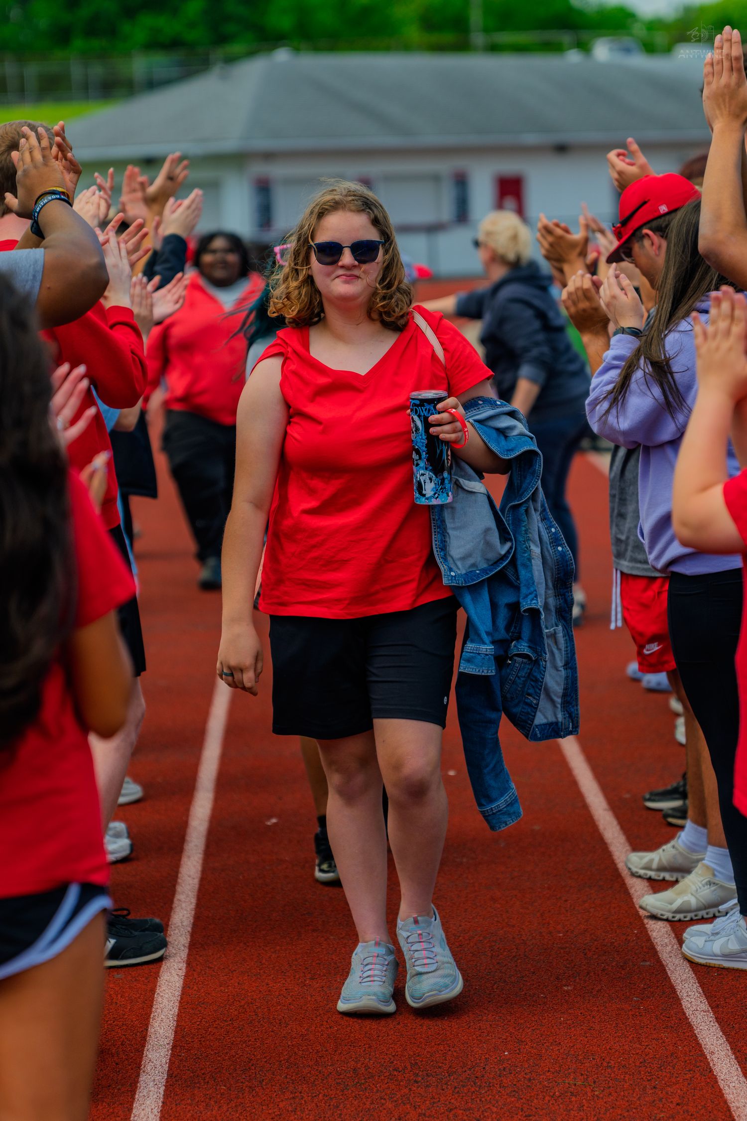 Young woman in red top walks through cheering crowd on a red running track during a school sports event.