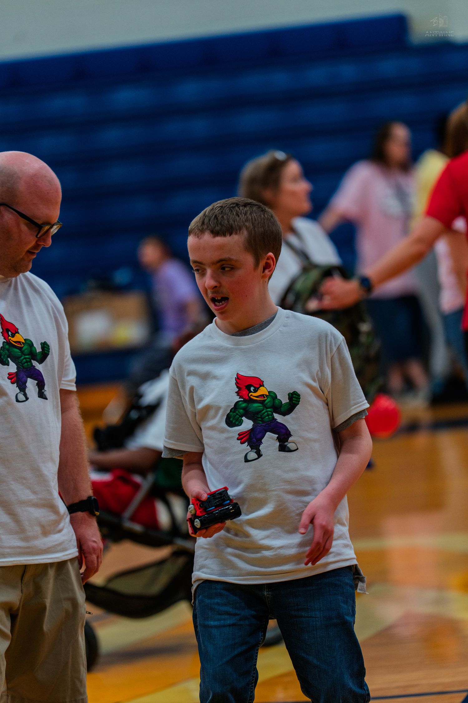 Boy in colorful cardinal mascot t-shirt holds remote control at indoor school gymnasium event.