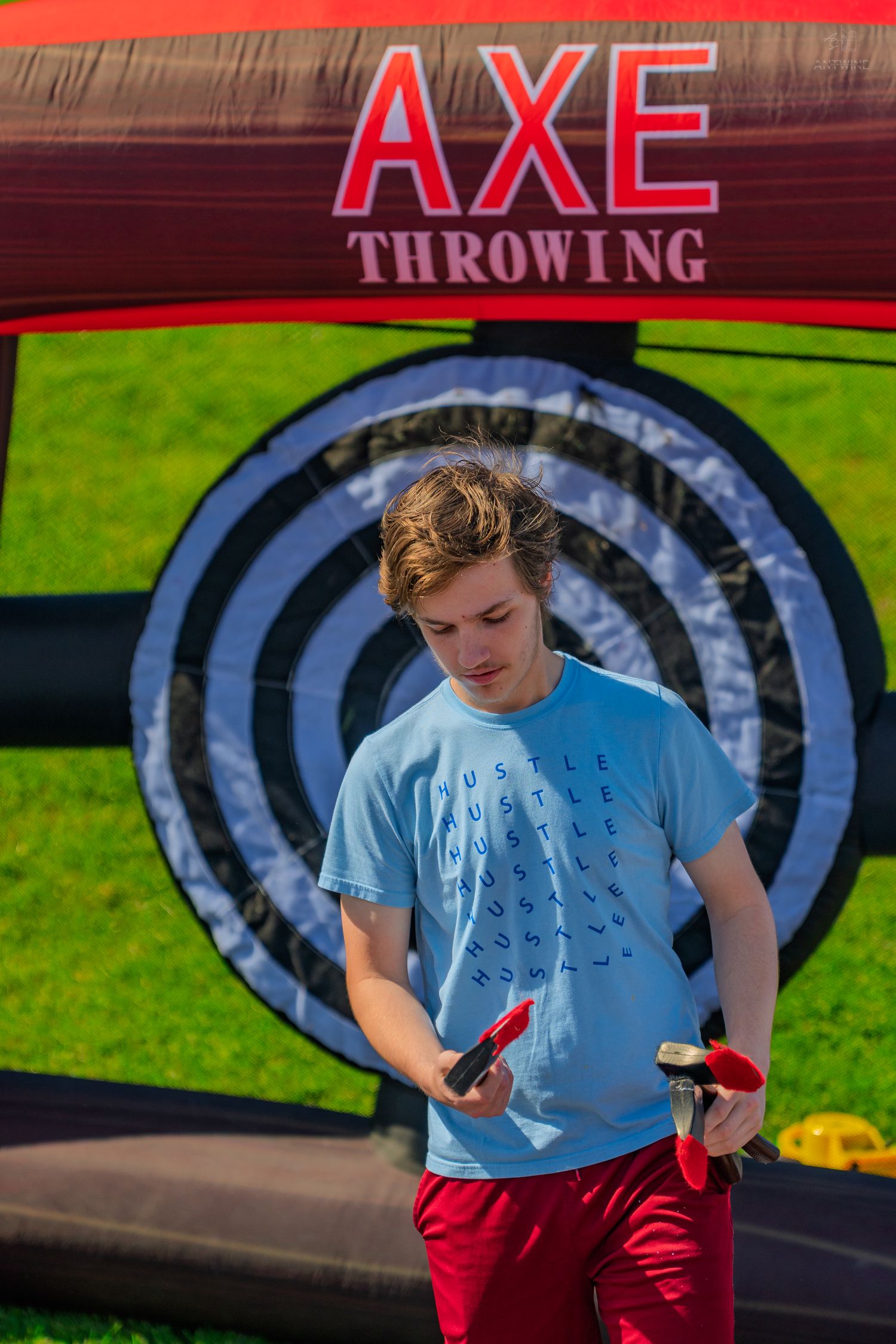 Teen boy holding axes at an inflatable axe throwing target game outdoors on a sunny day.