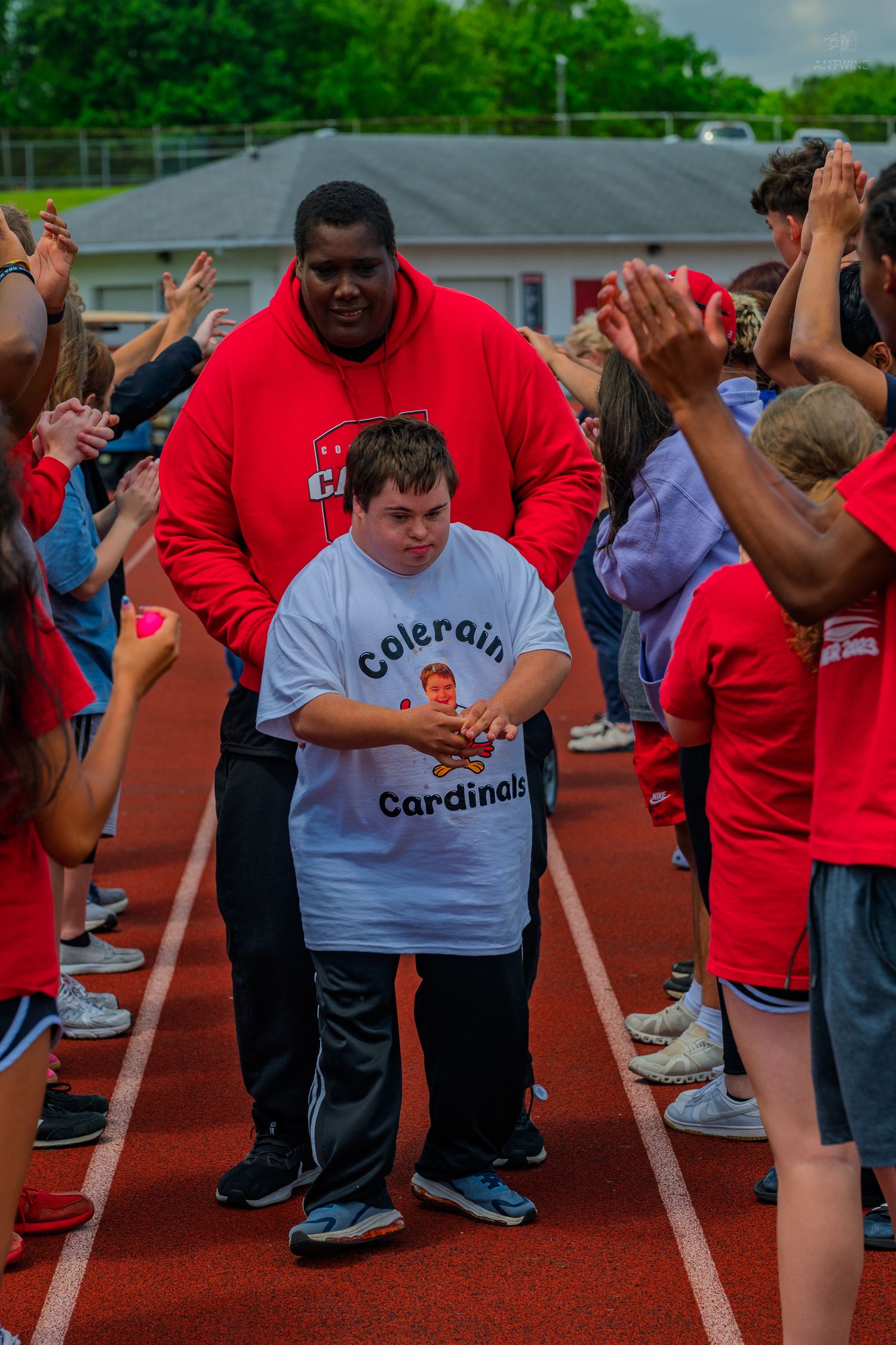 Student in Colerain Cardinals shirt walks track flanked by cheering peers and adult supporter at school sports event.
