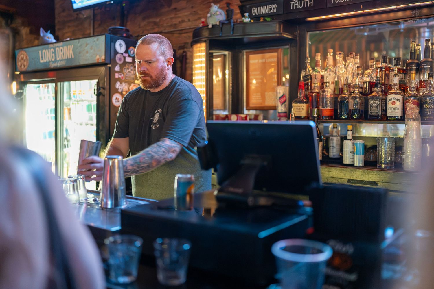 Tattooed male bartender mixing drinks behind a well-stocked bar with bottles and tap handles.