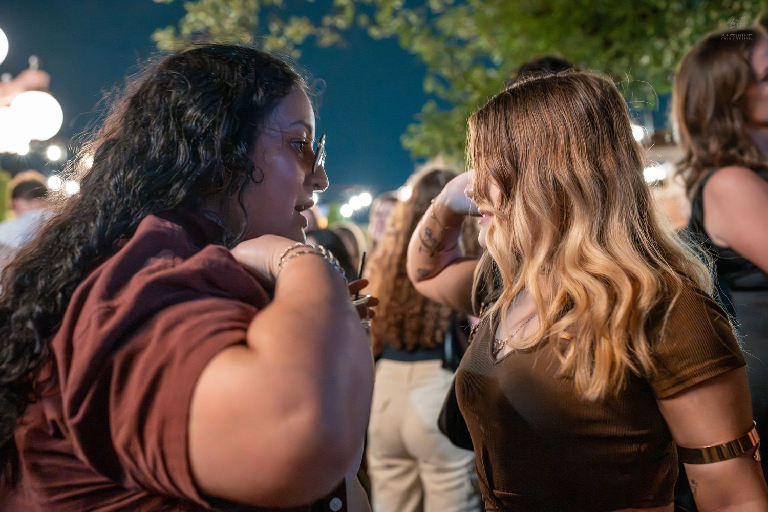 Two women having a conversation at an outdoor evening event with crowds in the background.