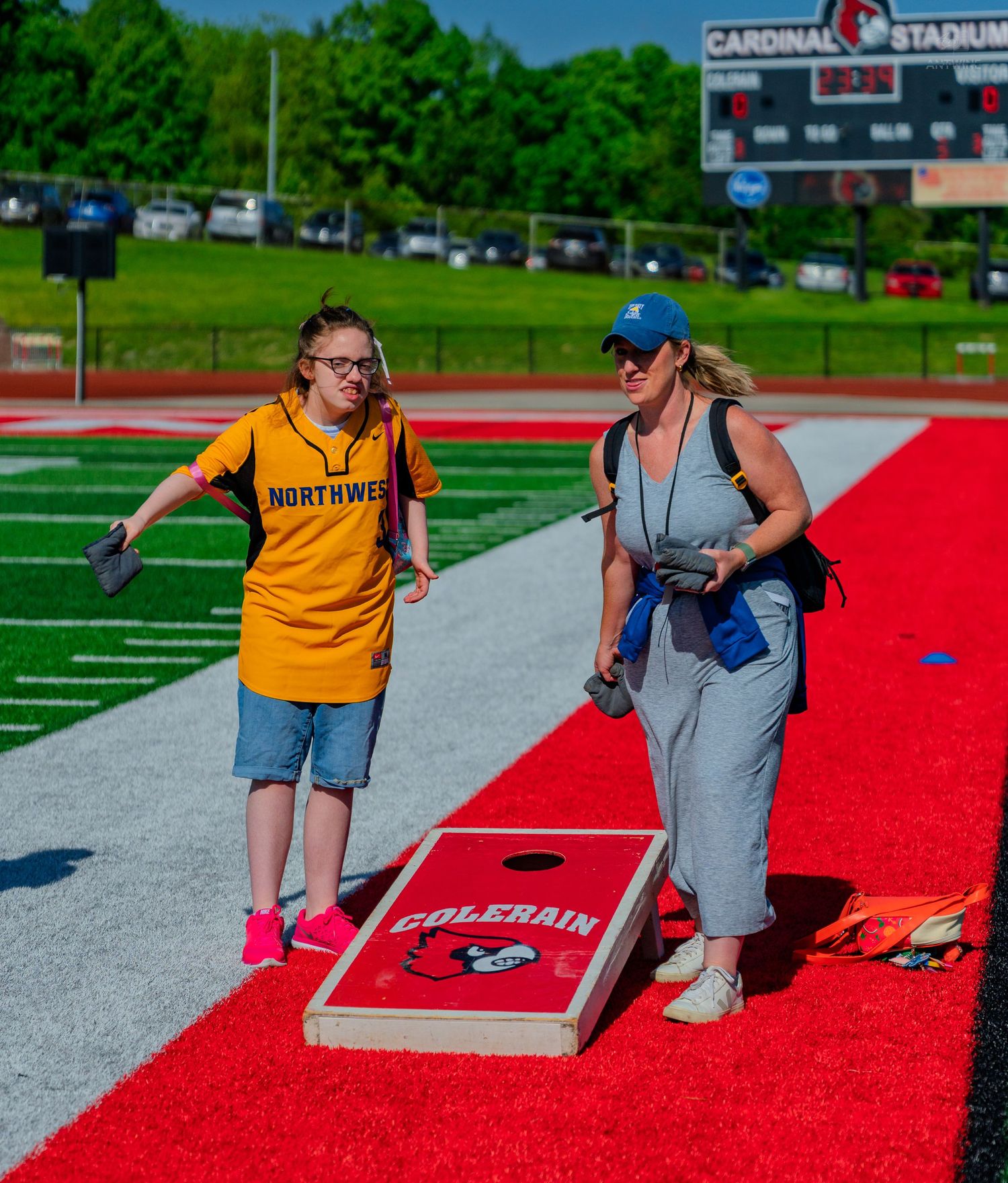 Two women playing cornhole on a red track at Cardinal Stadium, one wearing a Northwest jersey.
