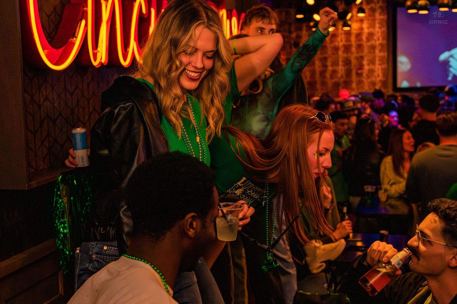 Women in green outfits dancing and celebrating under neon Cincy Love sign at a bar party.