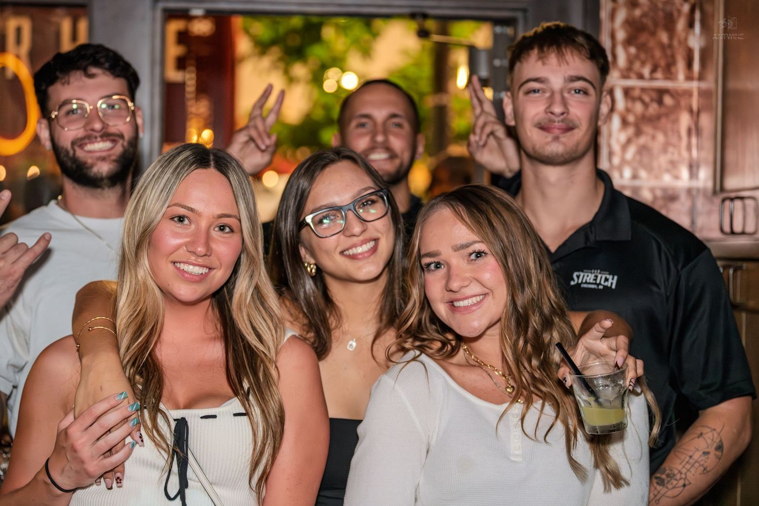Three young women in white tops smiling with friends at a lively bar event.