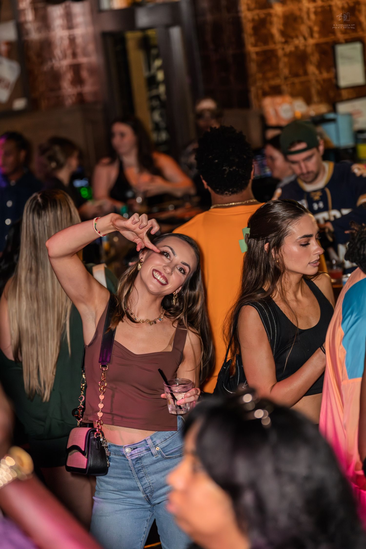 Smiling woman in brown crop top holding a drink poses at a lively bar while friends socialize in the background.