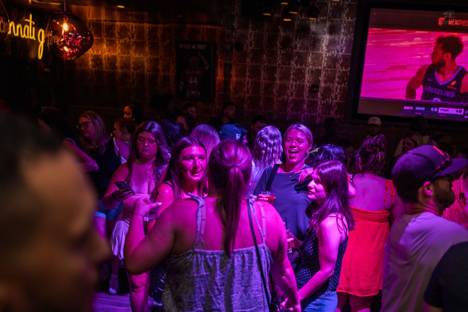 People dancing under pink and purple lights in a lively nightclub with a screen in background.