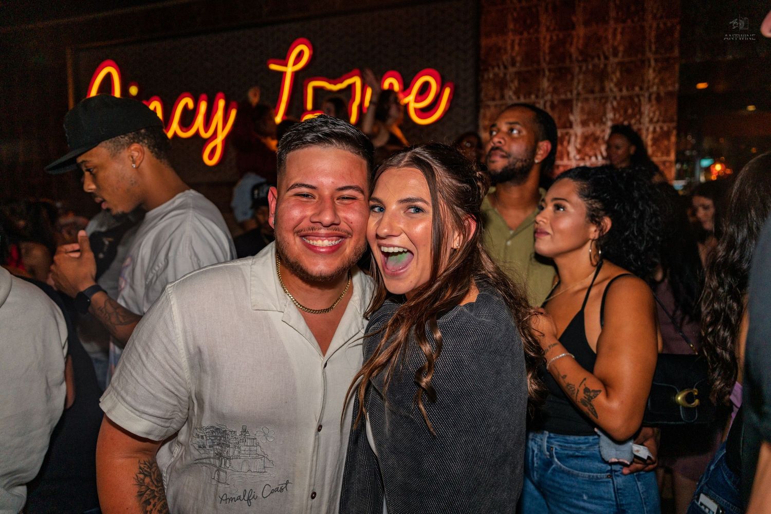 Smiling couple posing together at a lively nightclub with neon sign in the background.