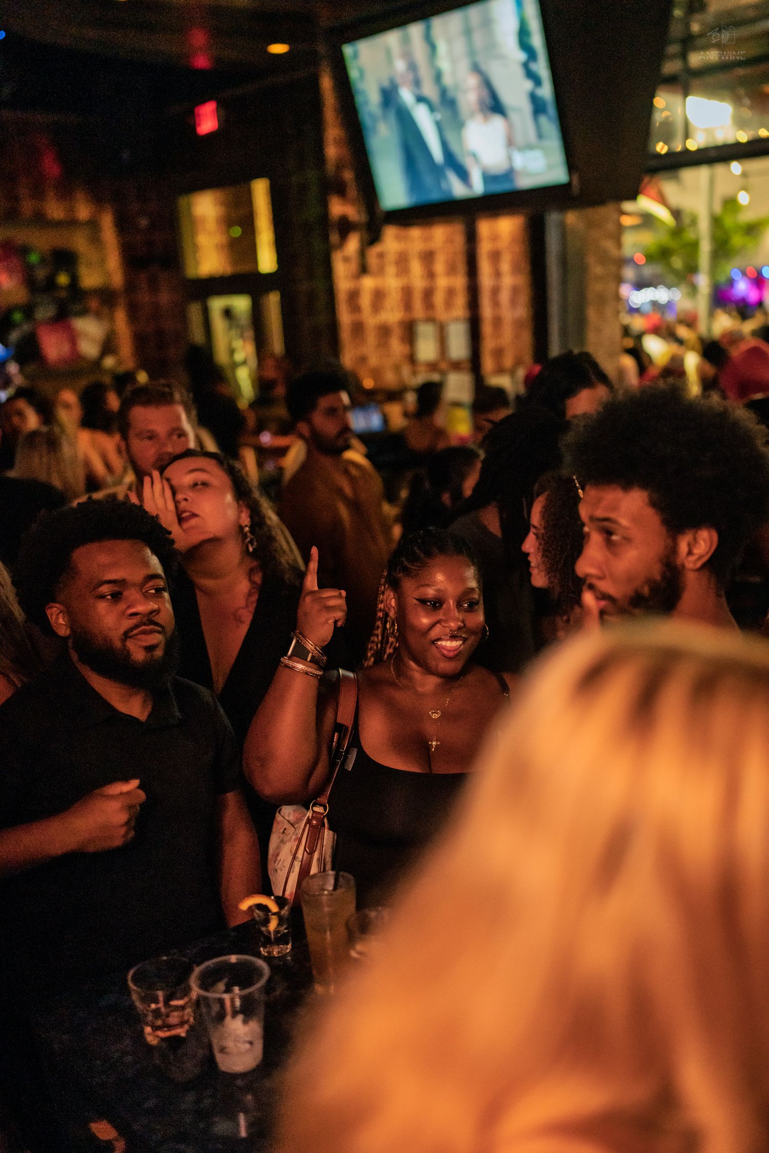 Diverse group of friends laughing and pointing inside a lively bar with TV screens above.