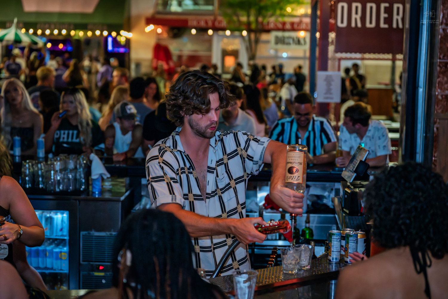 A bartender in a patterned shirt pours drinks at a busy outdoor bar venue at night.