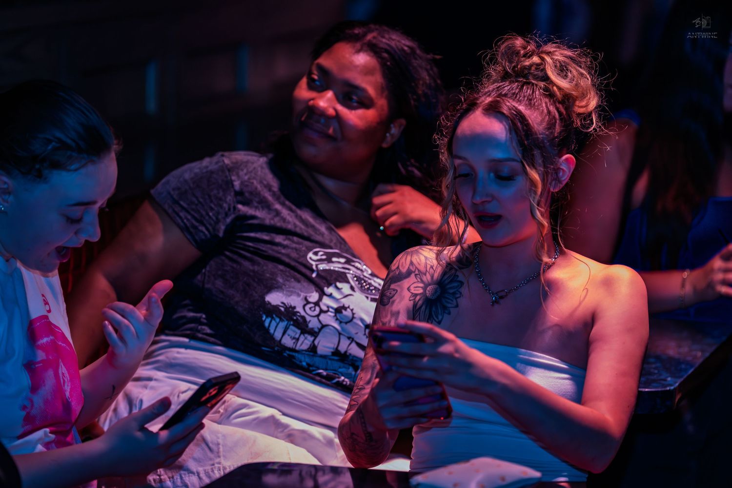 Three women looking at smartphones together in a dimly lit nightclub setting.