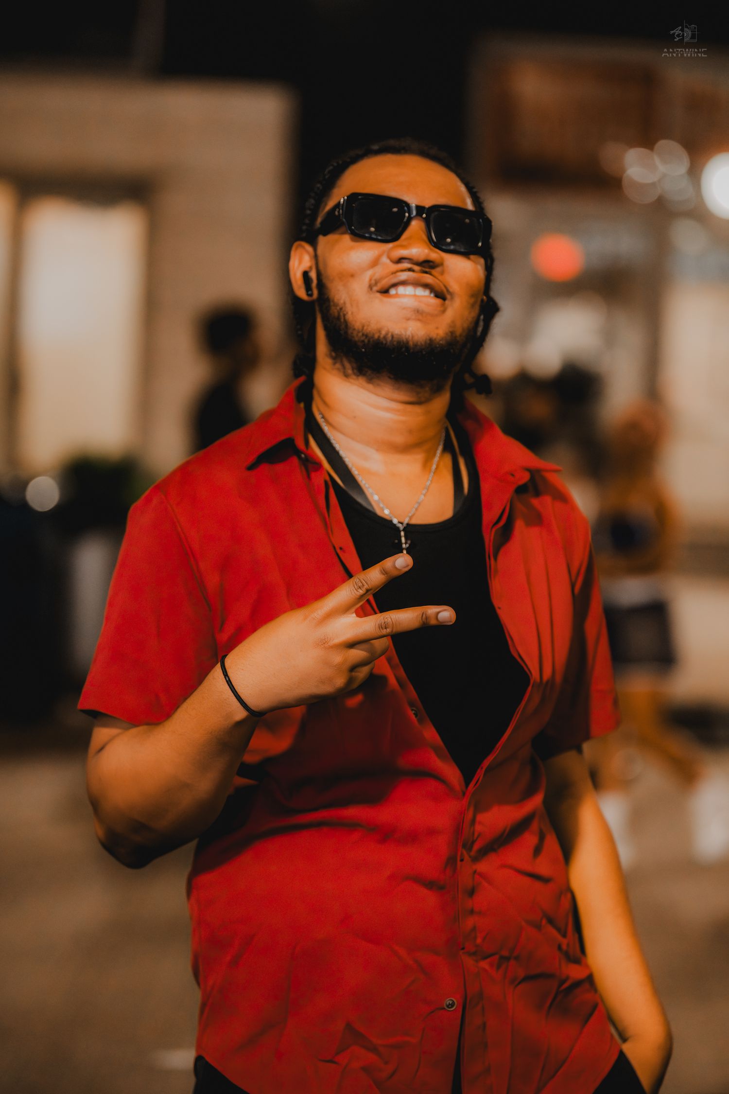A man in a red shirt and sunglasses smiles while making a peace sign at an outdoor nighttime event.