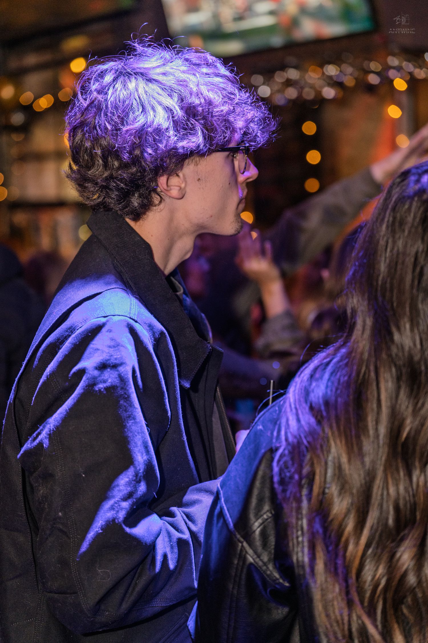 Young man with curly hair and glasses dancing under purple lights at a nightclub party.