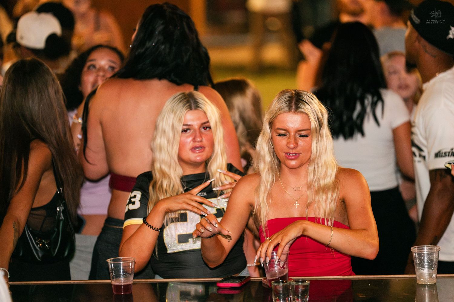 Two blonde women standing at a nightclub bar counter with drinks, looking at phones.