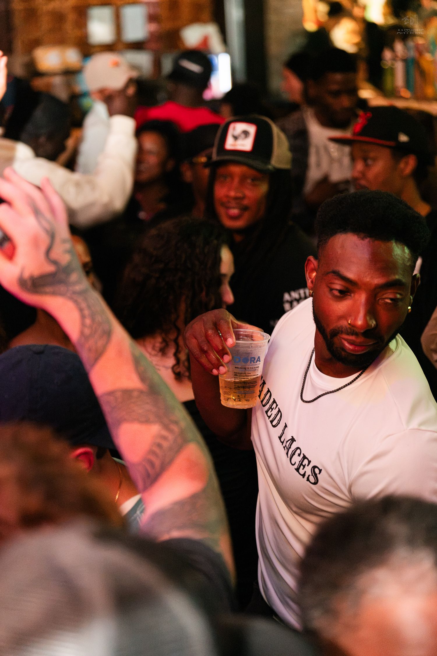 A man in a white t-shirt holds a drink while navigating a crowded bar event.