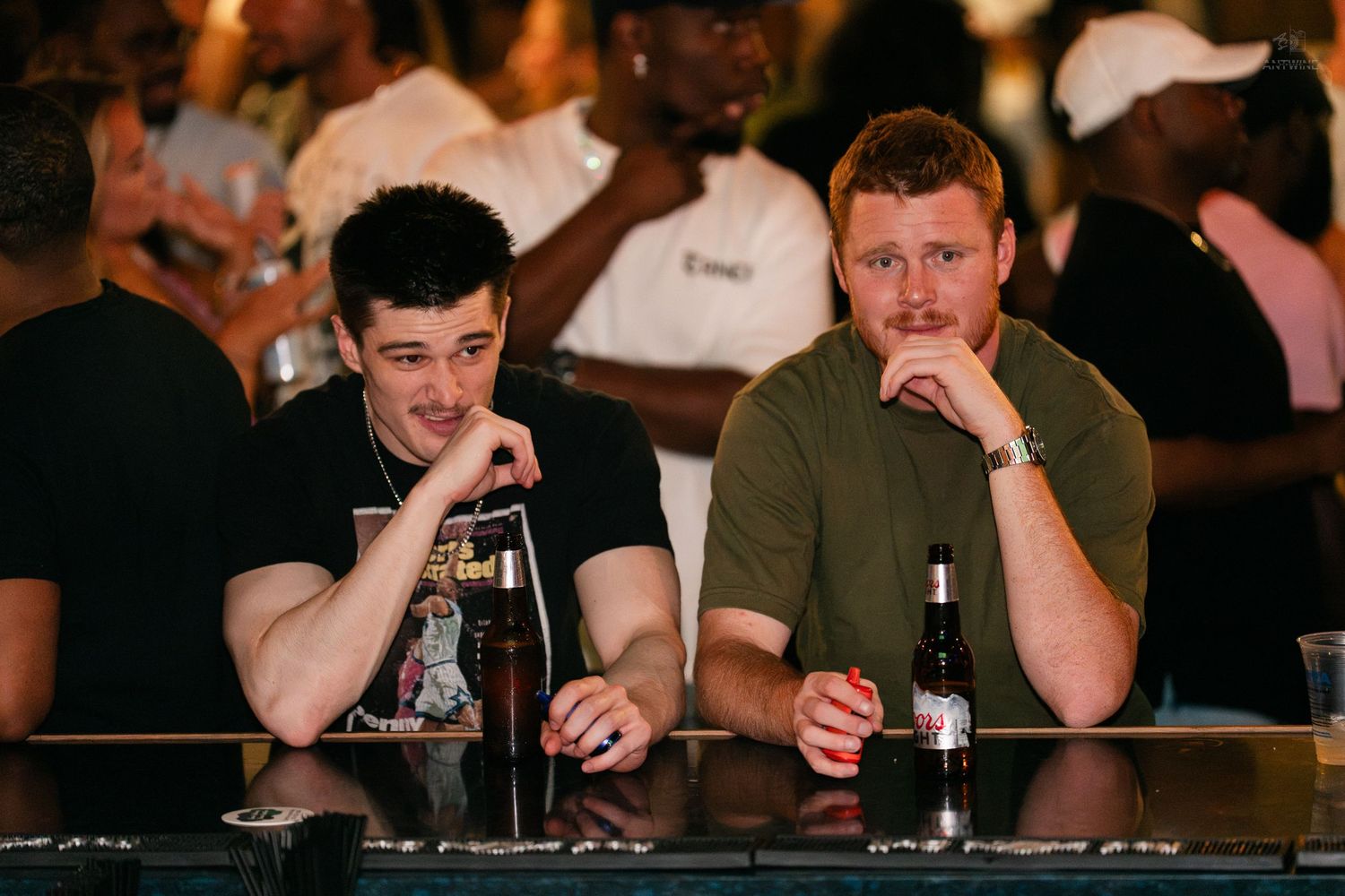 Two young men sitting at a bar counter with beer bottles.