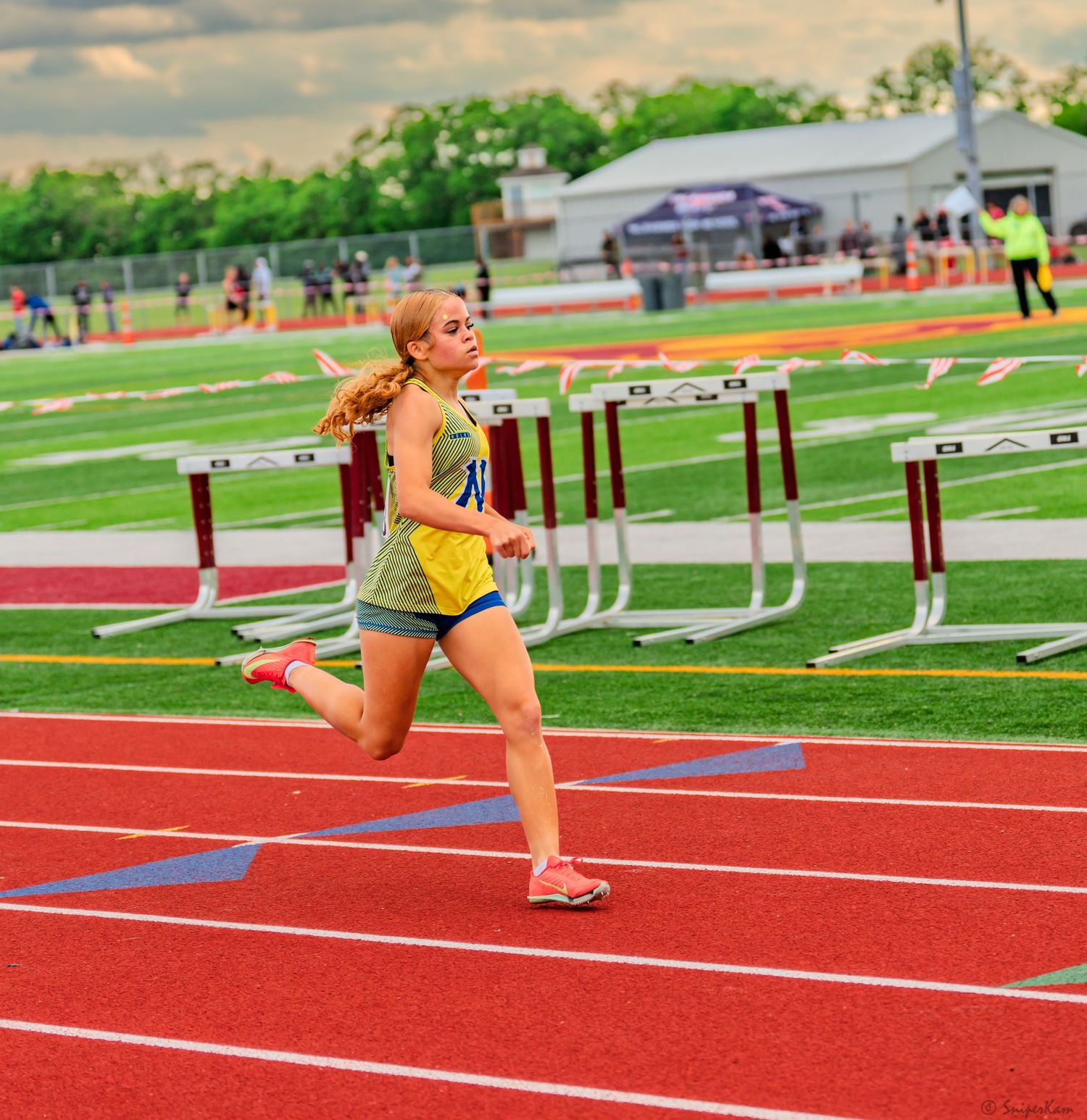 Young female athlete in yellow uniform sprints past hurdles on a red competition track.