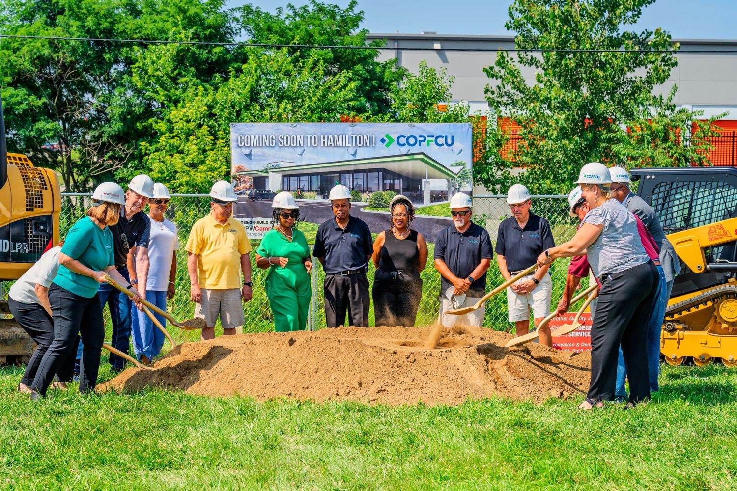 Large group celebrating COPFCU groundbreaking ceremony with shovels in sunny outdoor setting.