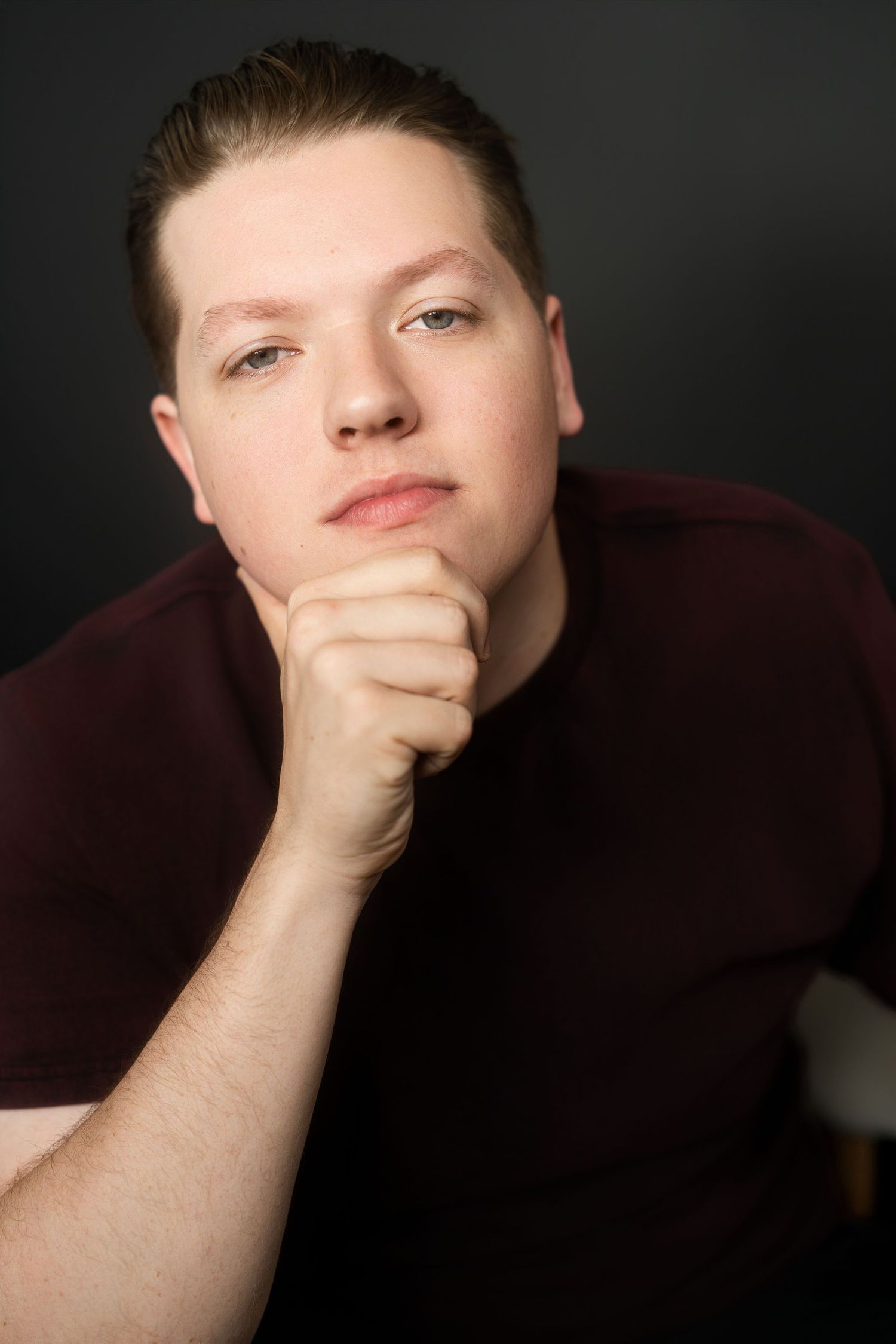Studio portrait of young man in maroon shirt with fist under chin against dark gray background.