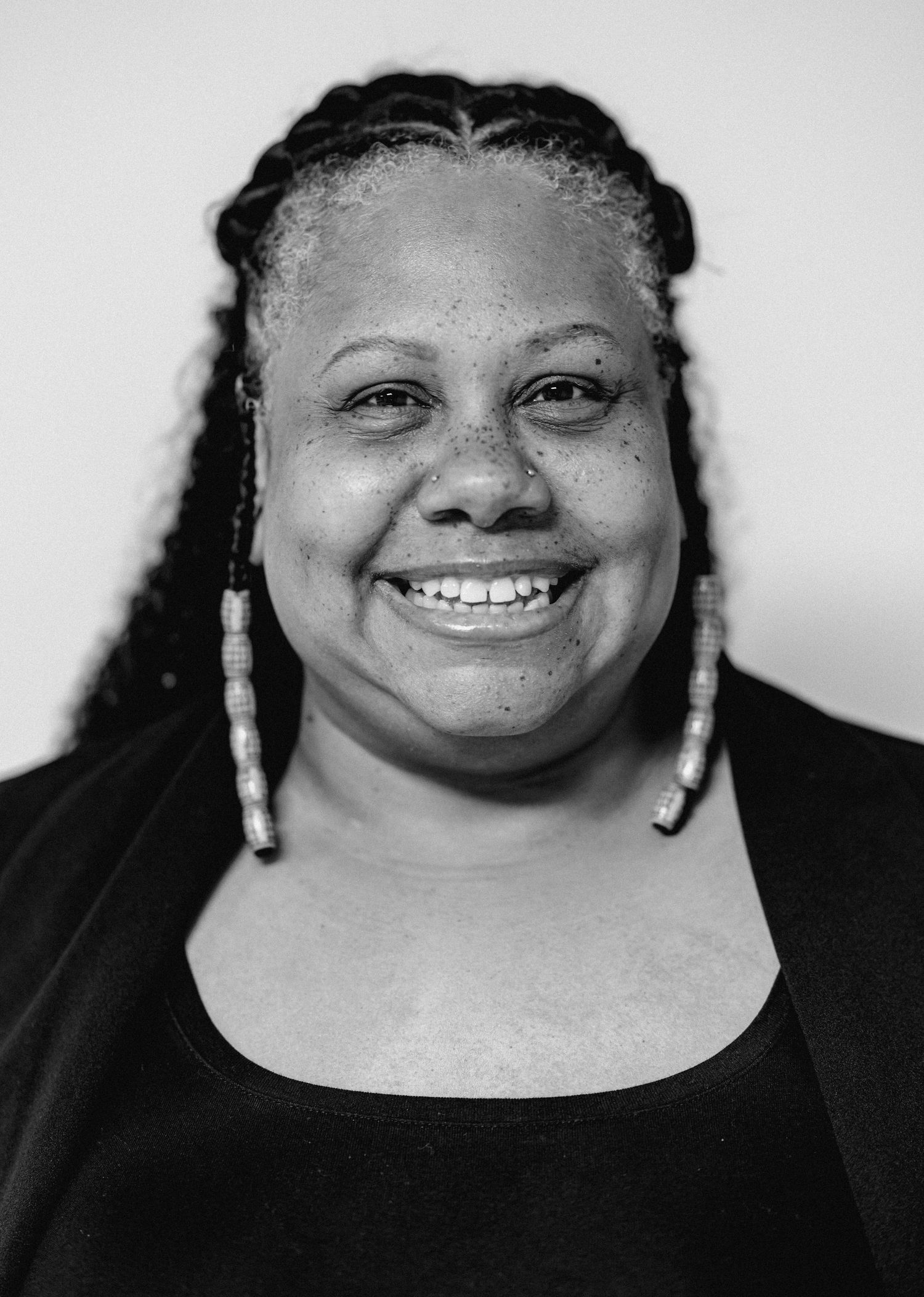 Smiling Black woman with braided hair and beaded earrings wearing black top, black and white portrait photo.