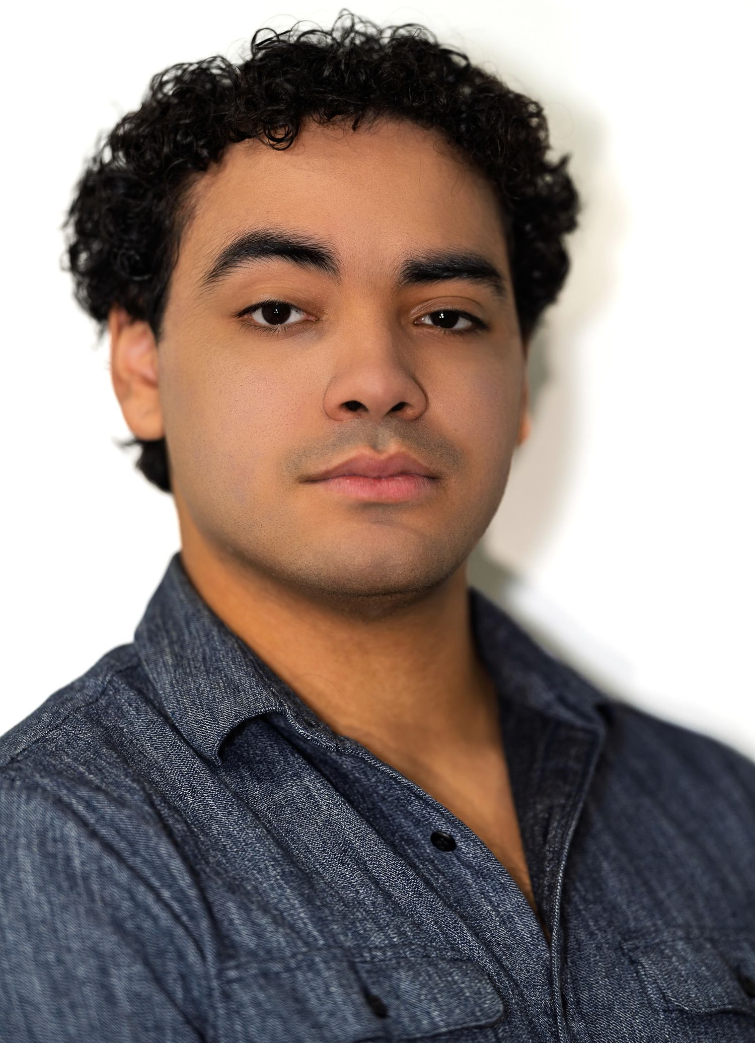 Young man with curly black hair wearing a blue denim shirt in a professional color headshot photo.