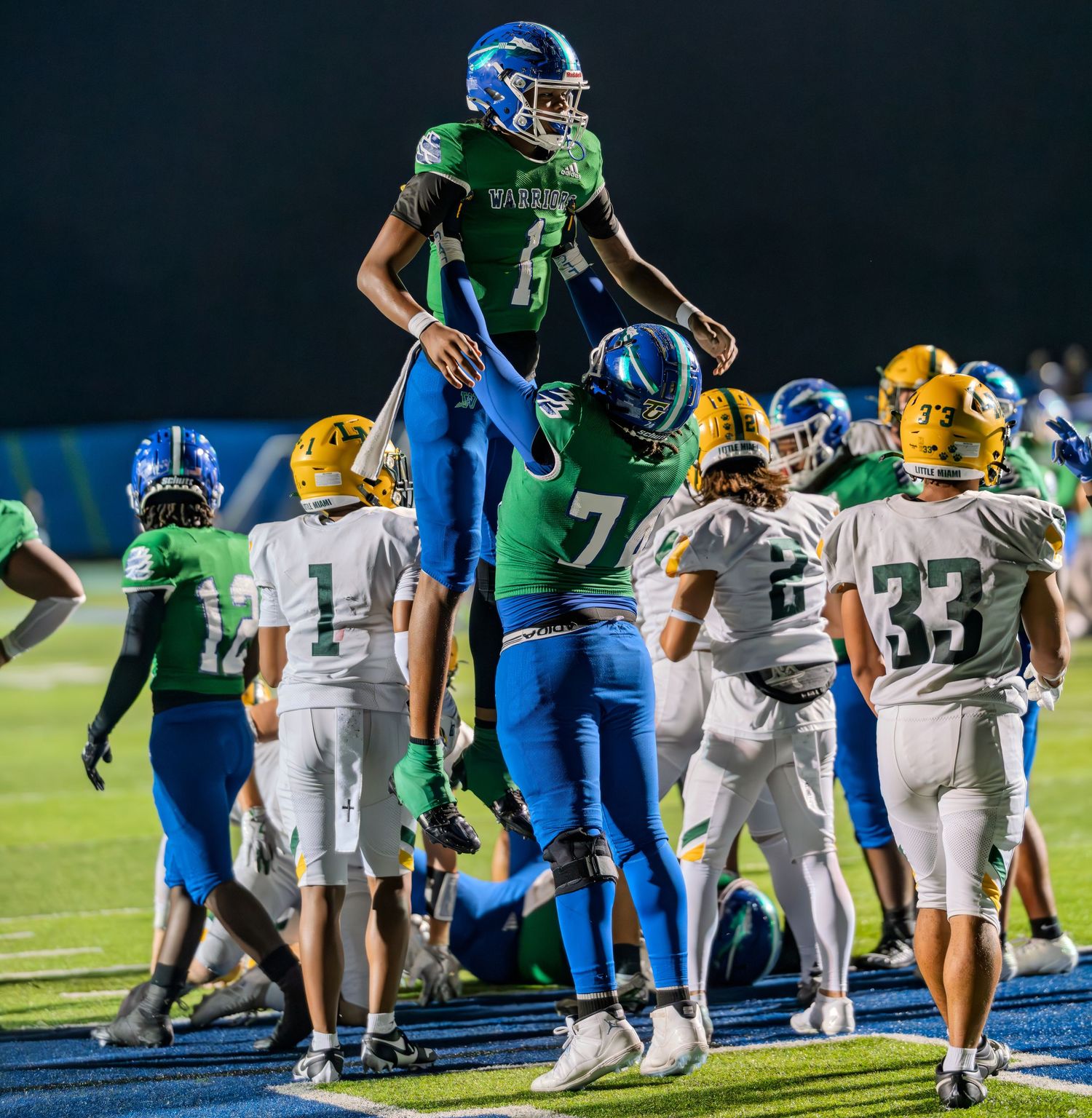 Two Warriors football players in green and blue uniforms celebrate a touchdown on the field.
