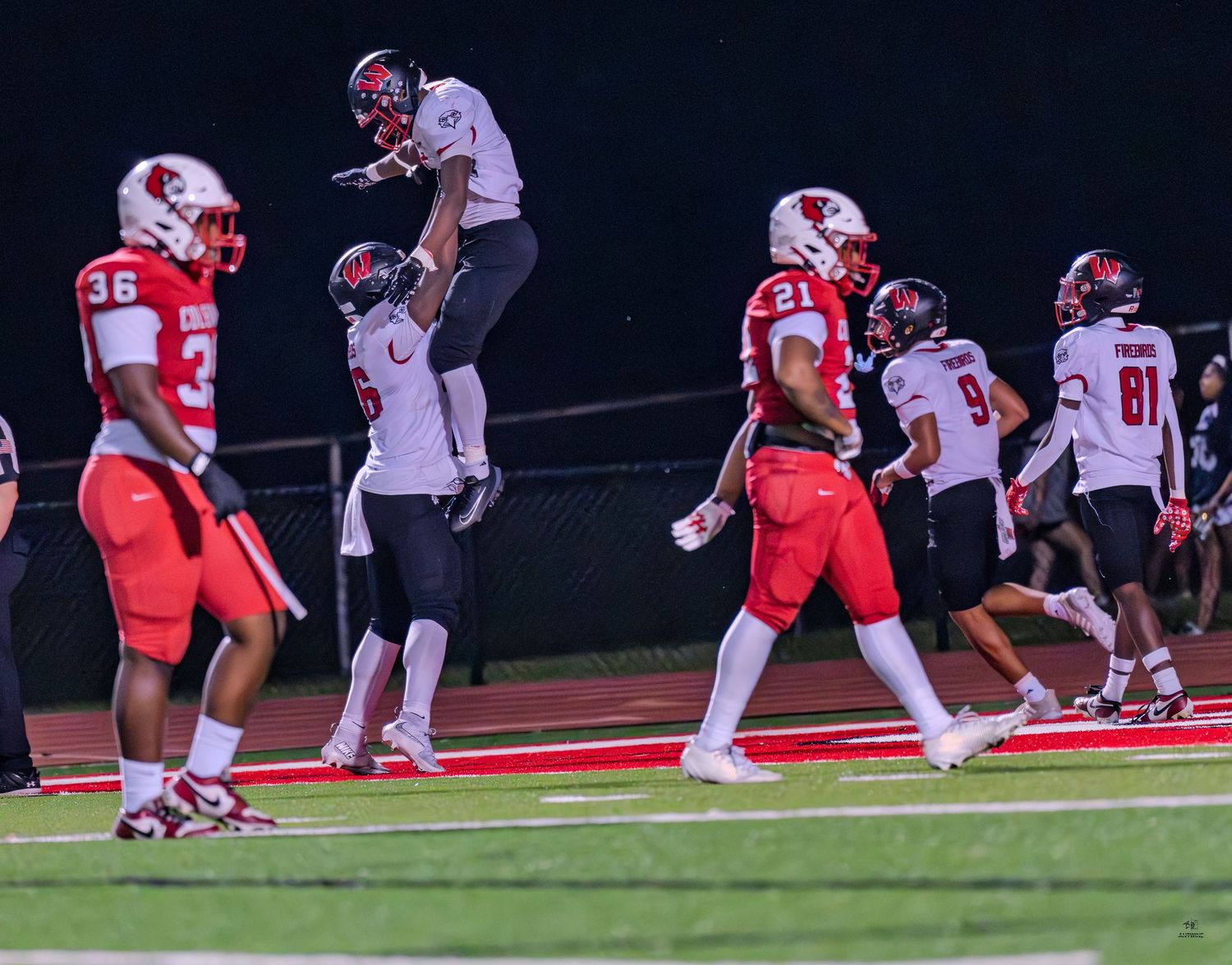 Football player lifted in celebration by teammate during a high school night game on a green field.