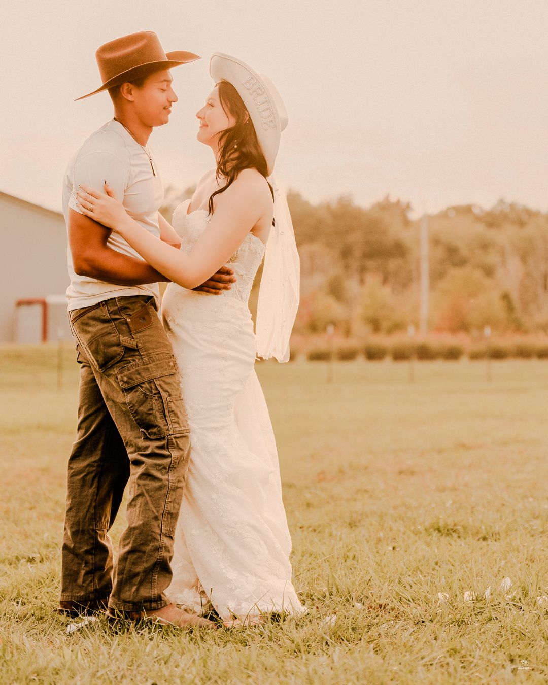 Bride in white gown and cowboy hat embracing groom in brown hat at golden hour on farm.