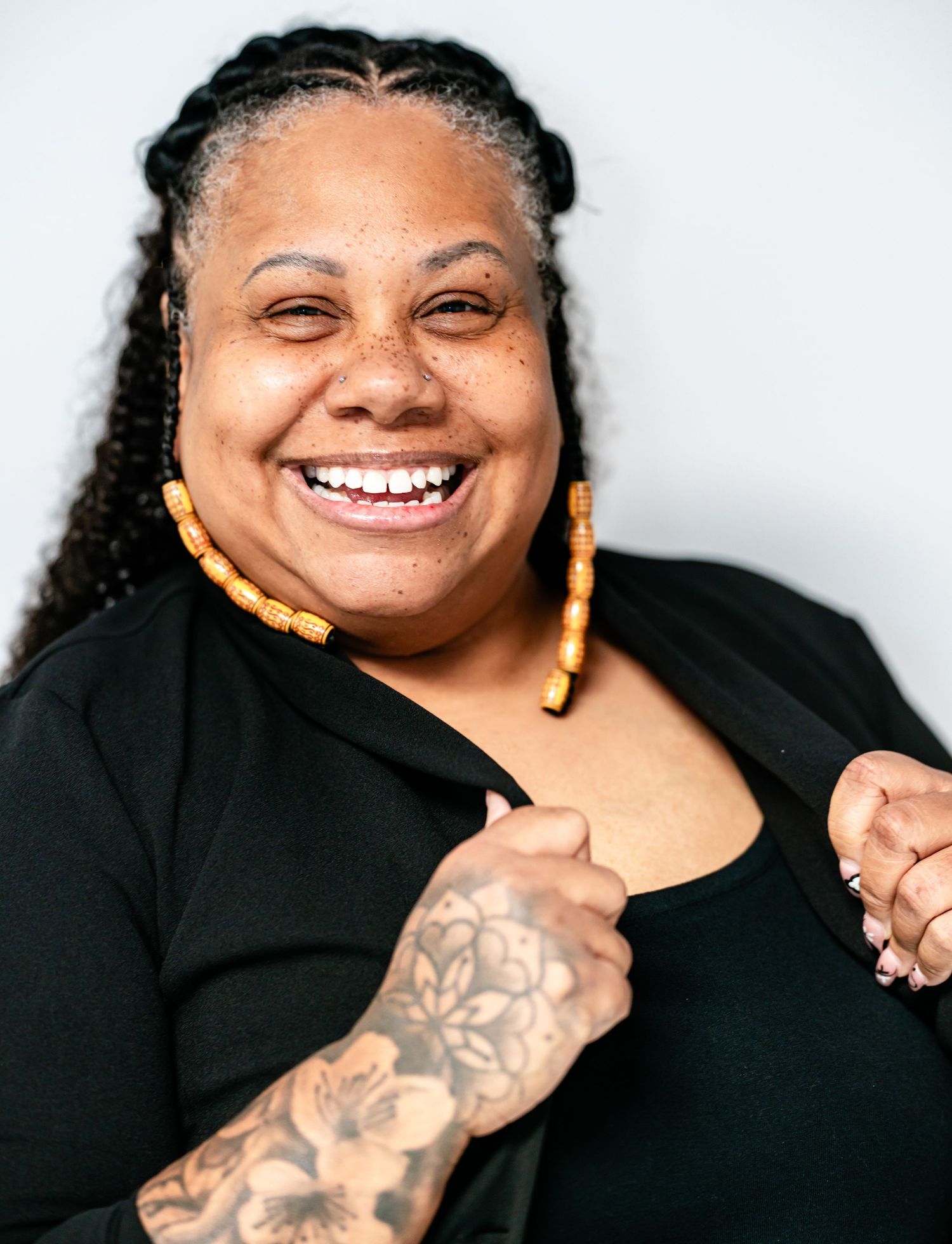 Laughing Black woman with braided hair and yellow earrings showing floral arm tattoo, candid portrait.