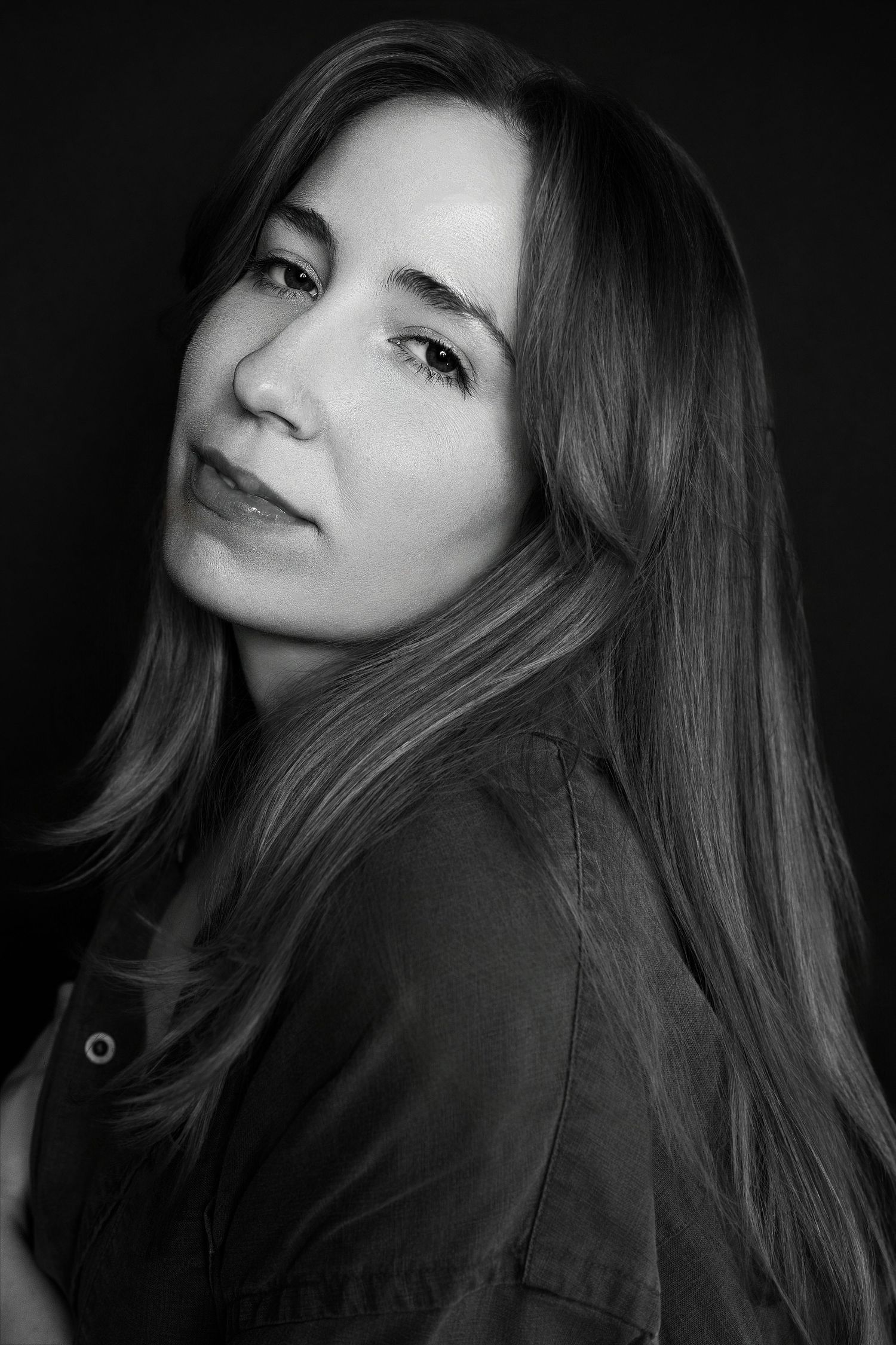 Black and white side portrait of young woman with long hair gazing over shoulder against dark background.