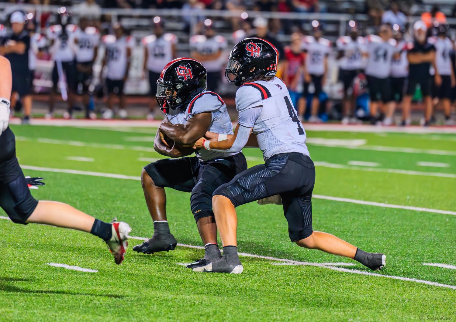Two football players execute a handoff play on a green field during a night game.