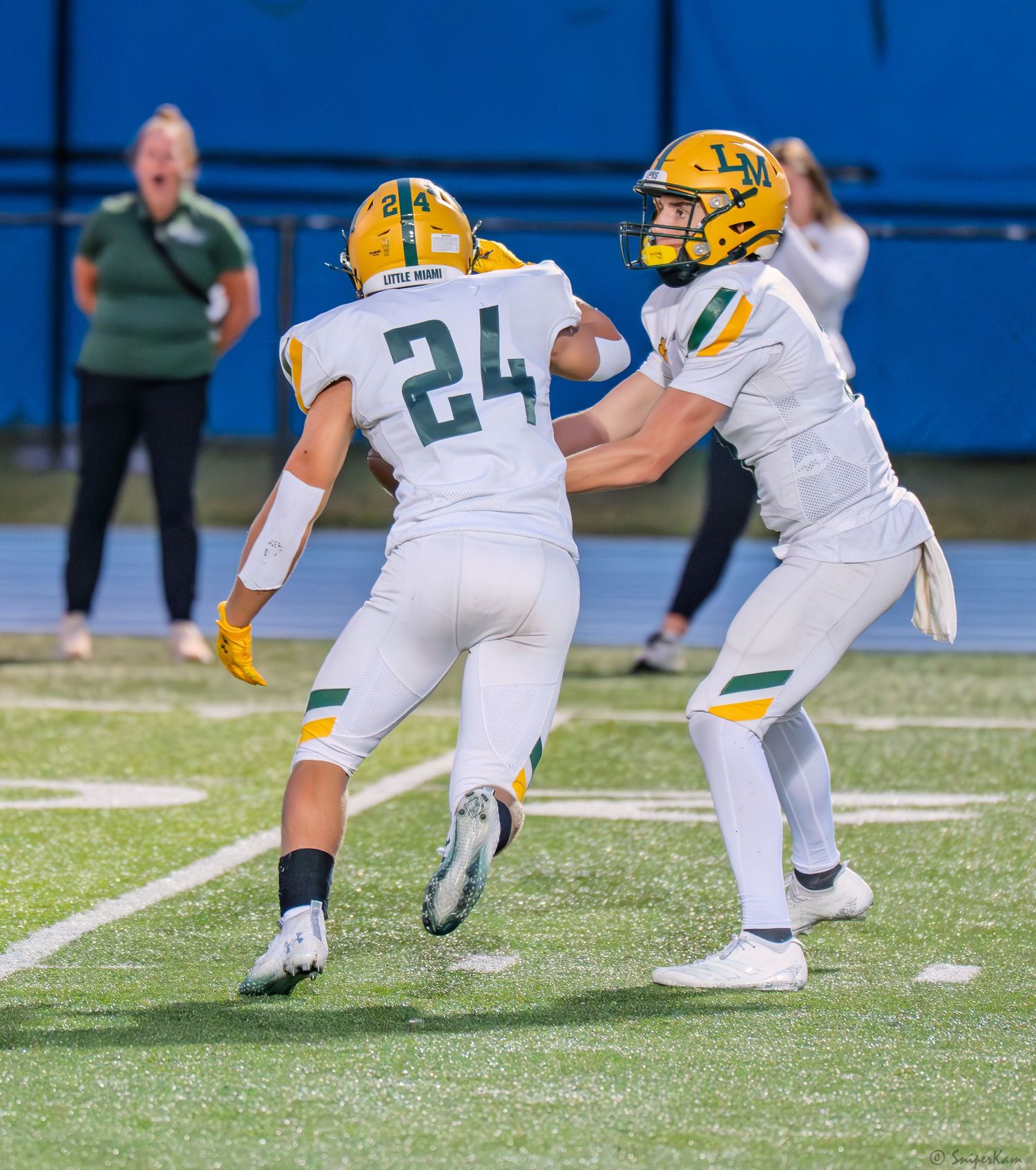 High school football player number 24 receives handoff from quarterback during night game on blue turf field.