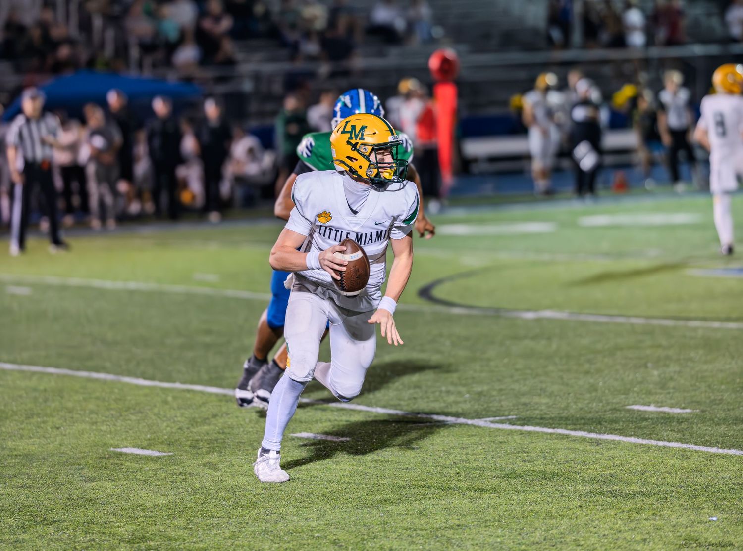 High school football quarterback in white and green uniform scrambles with ball during night game.
