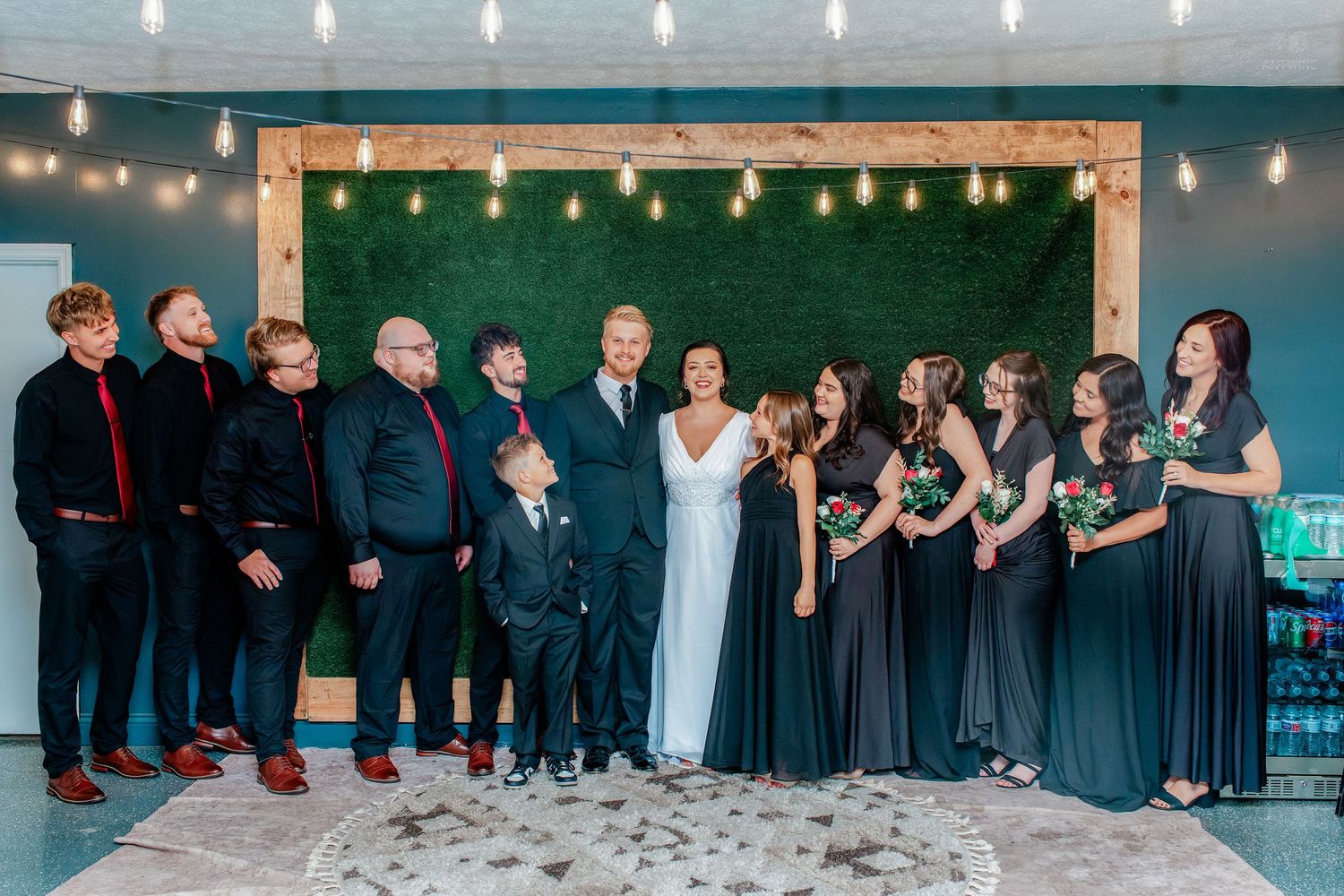 Wedding party poses together in front of a greenery wall with Edison bulb lights at an elegant indoor venue.
