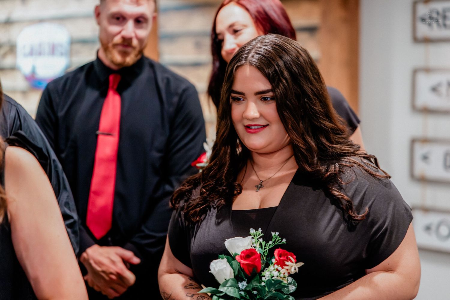 Woman in black dress holding red and white roses bouquet at a wedding ceremony with guests in background.