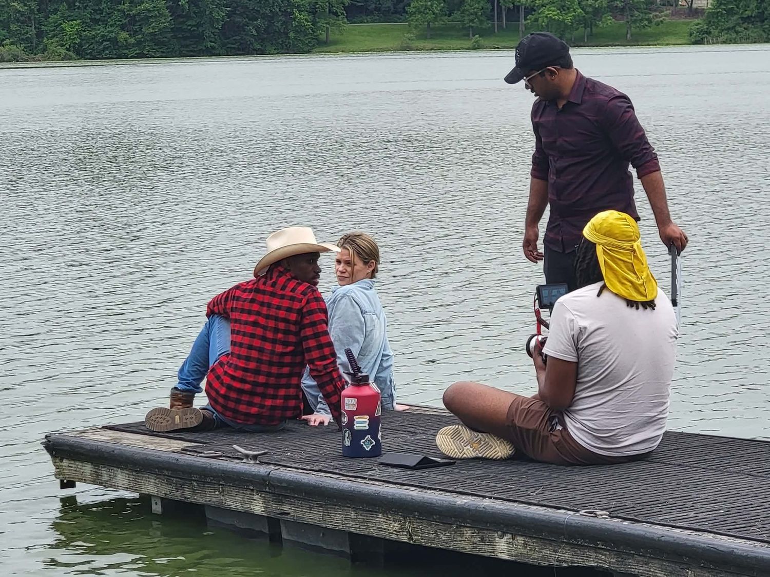 Group of people sitting on a wooden dock by a calm green lake, enjoying a relaxing outdoor fishing day together.