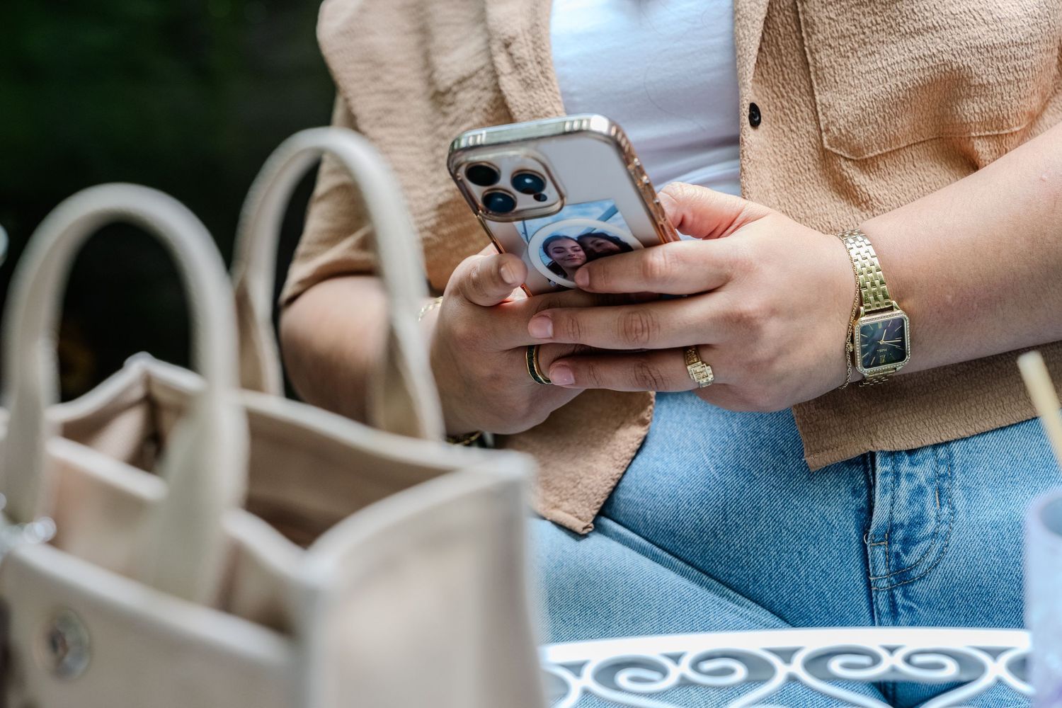 Close-up of woman holding iPhone with designer case beside beige tote bag outdoors.