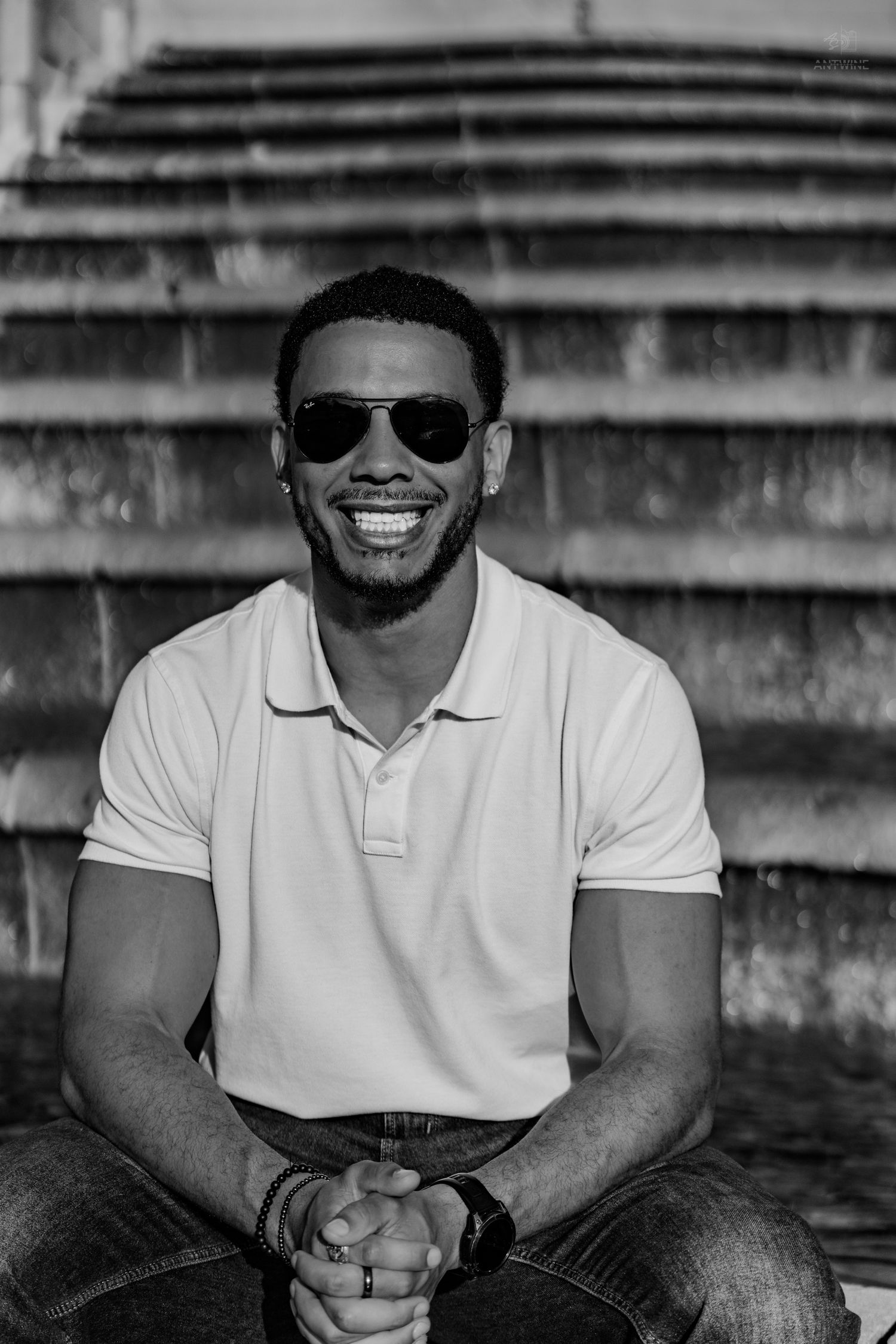 Black and white photo of a smiling young man in aviator sunglasses and polo shirt seated on steps.