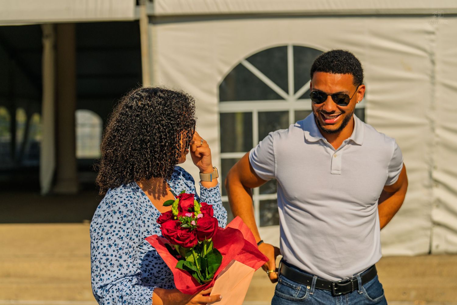A man in sunglasses smiles while presenting red roses to a woman in a blue floral dress outdoors.