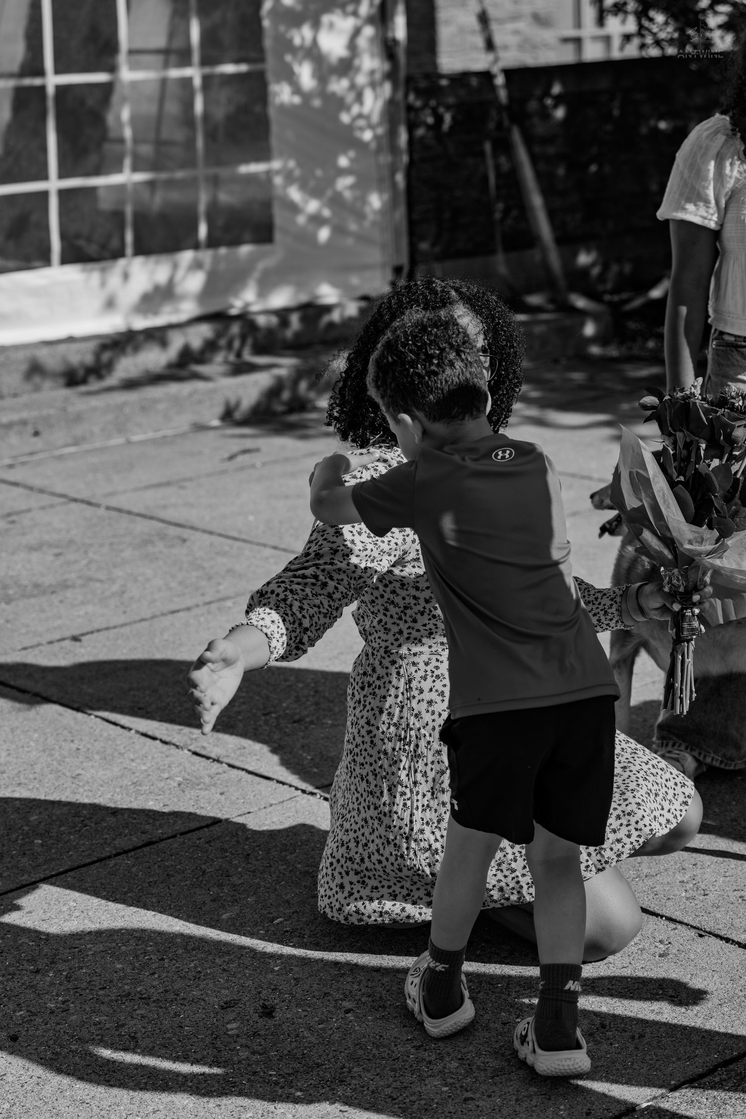 A child hugs a woman in a floral dress outdoors in black and white photography.